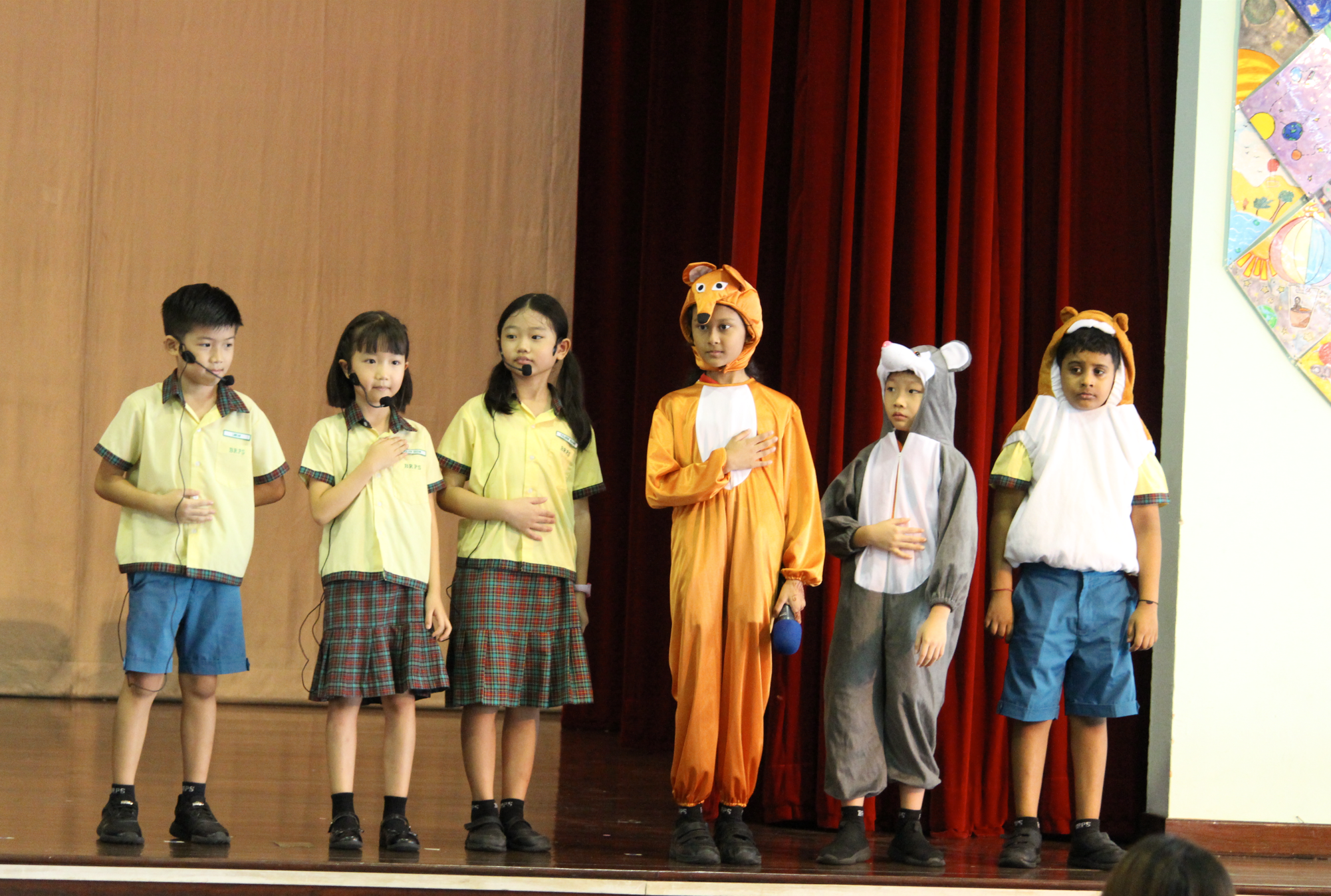 Six schoolchildren on a stage, three in uniforms and three in animal costumes, standing before red curtains.