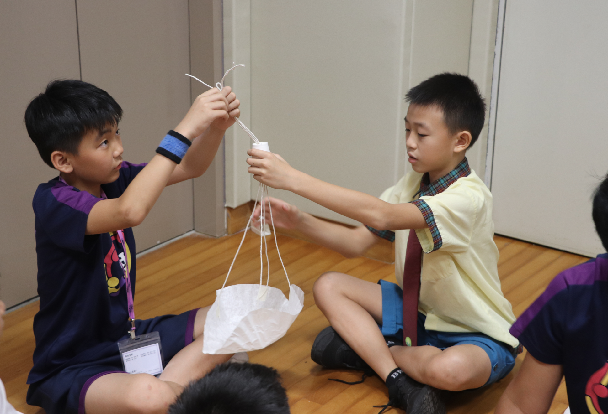 Two boys sit on a wooden floor, together assembling a small parachute made of white plastic bag and string.