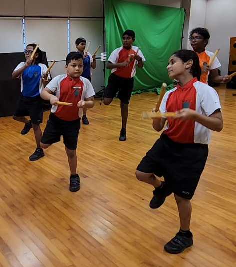 Six school children in red and white uniforms practice a stick routine on a wooden floor indoors.