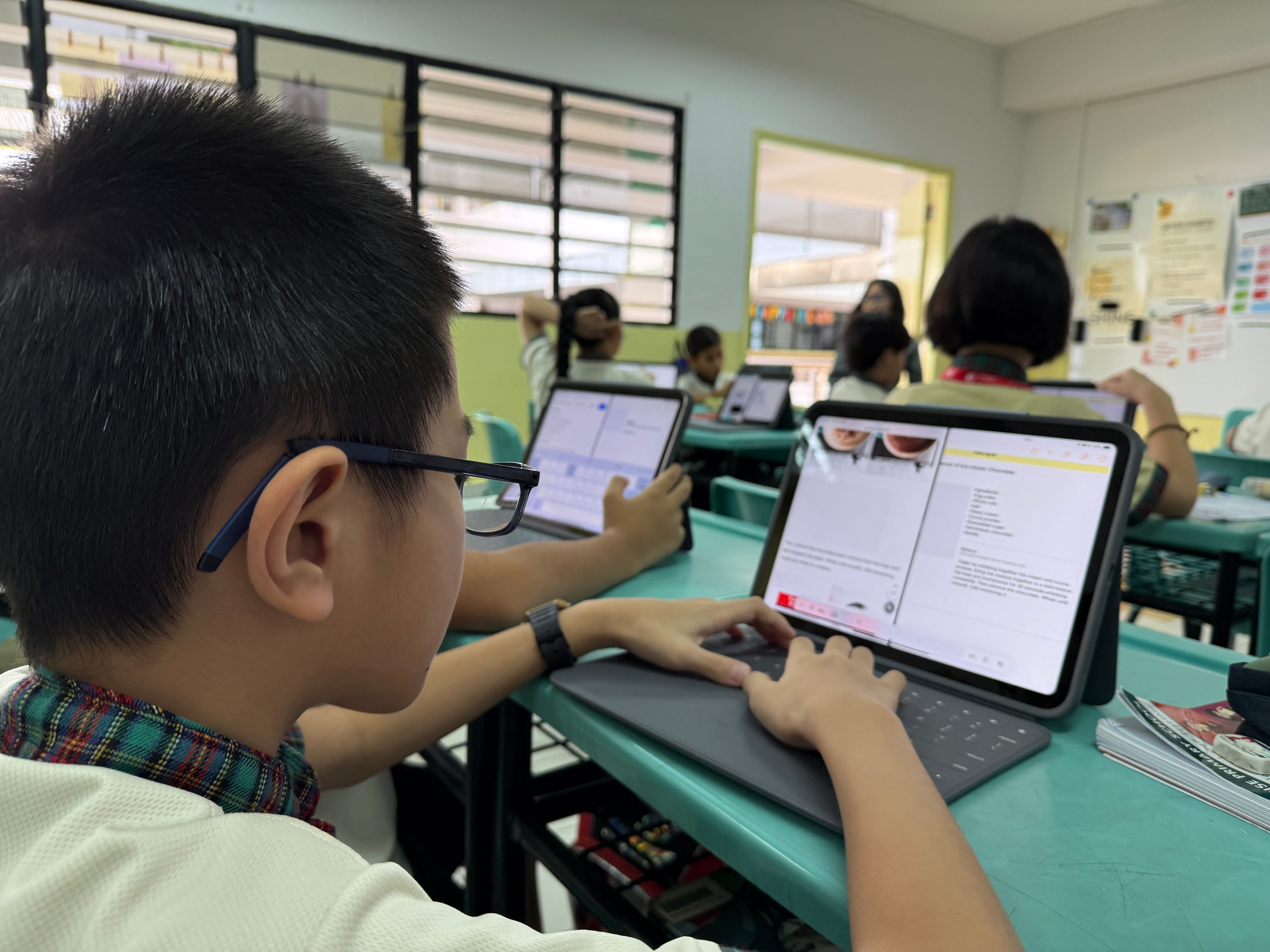 Student with glasses types on a tablet with keyboard at a classroom desk, other students in background.