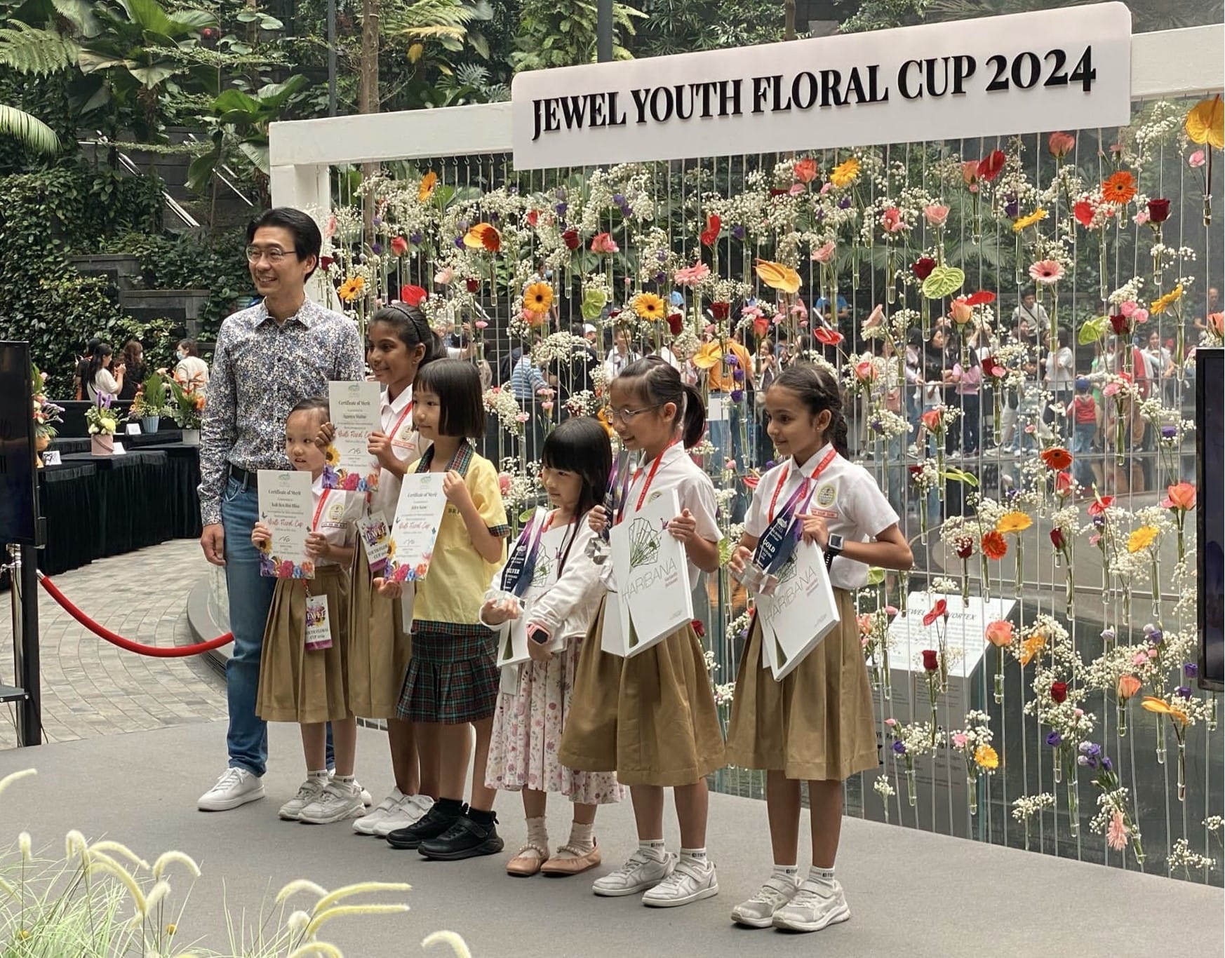 Children holding certificates and bags pose with an adult in front of a Jewel Youth Floral Cup 2024 floral display wall.