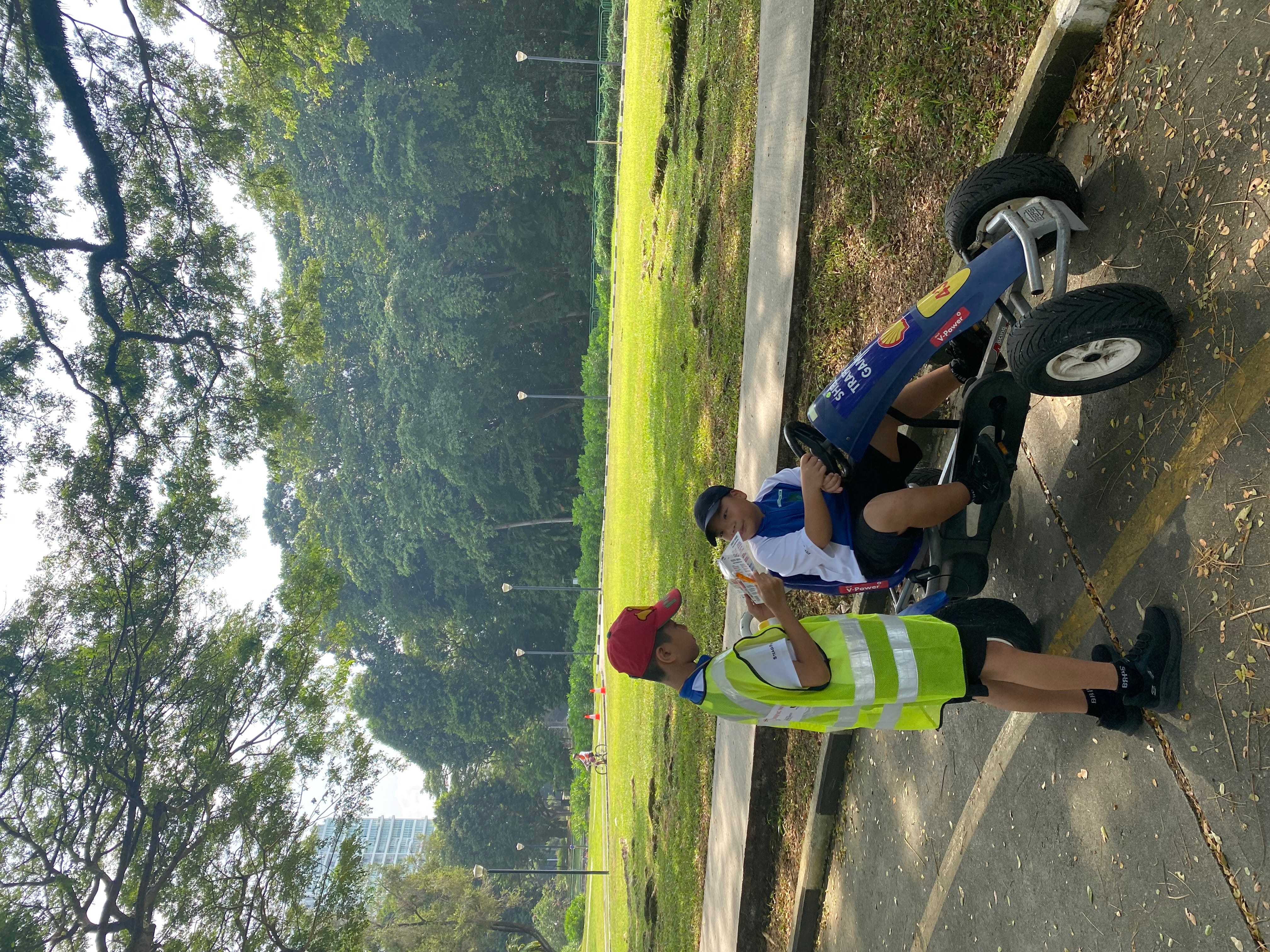 Child in a reflective vest gives a paper to a child seated in a pedal cart beside a park path.