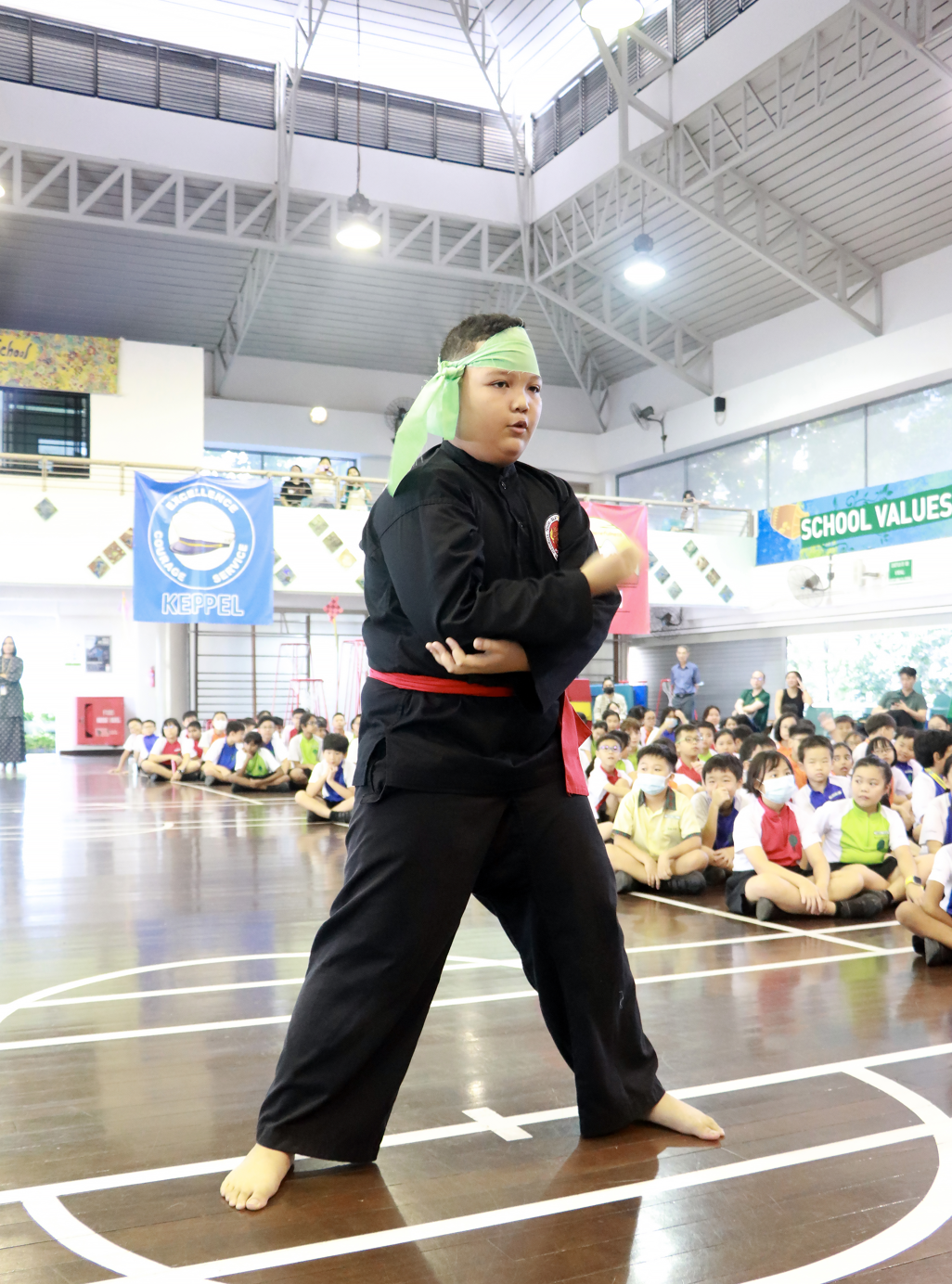 A child in black martial arts uniform with red belt and green headband performs in a school hall before seated students.