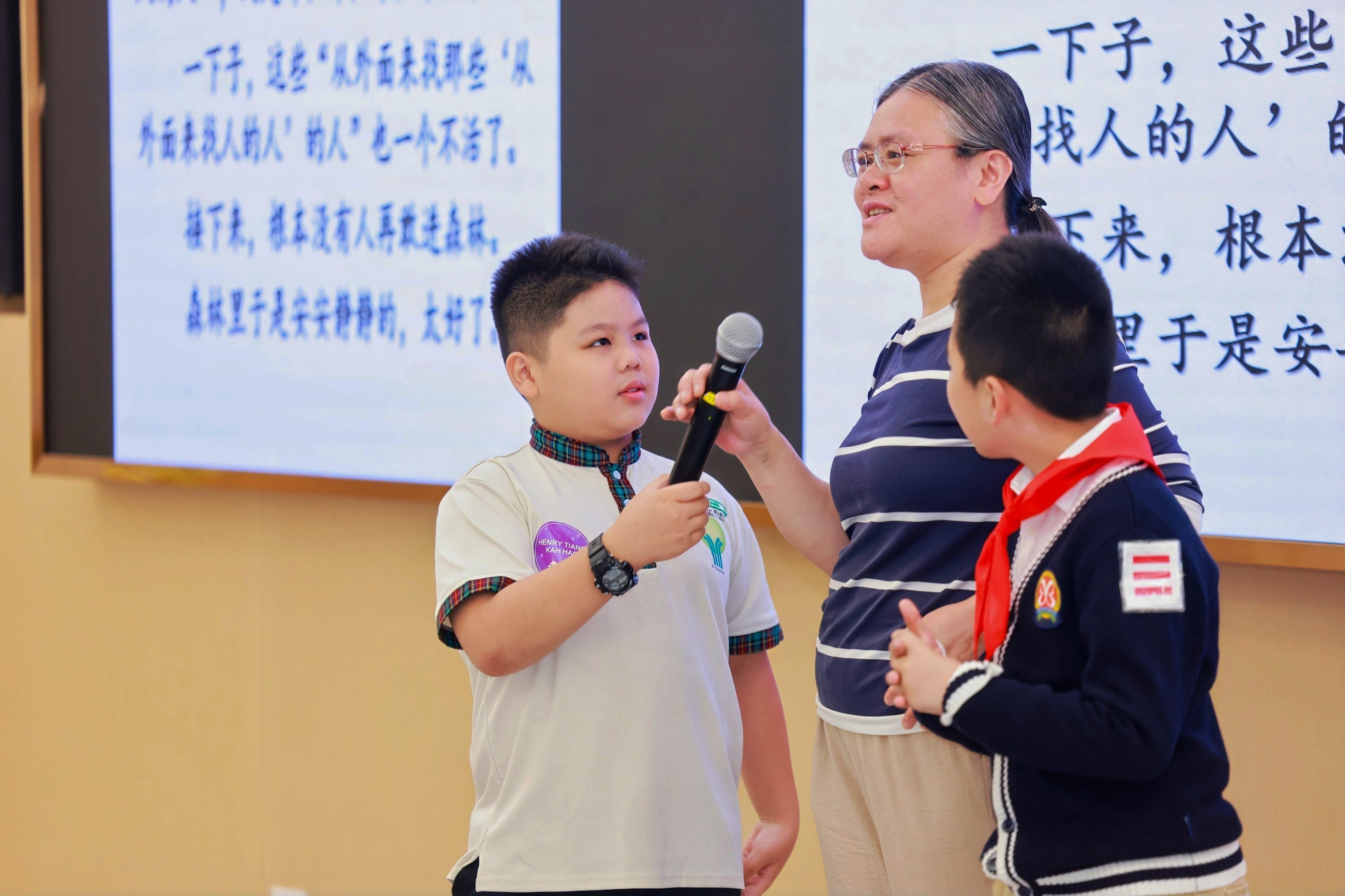 A boy holds a microphone while an adult and another child stand nearby in front of Chinese text on screens.