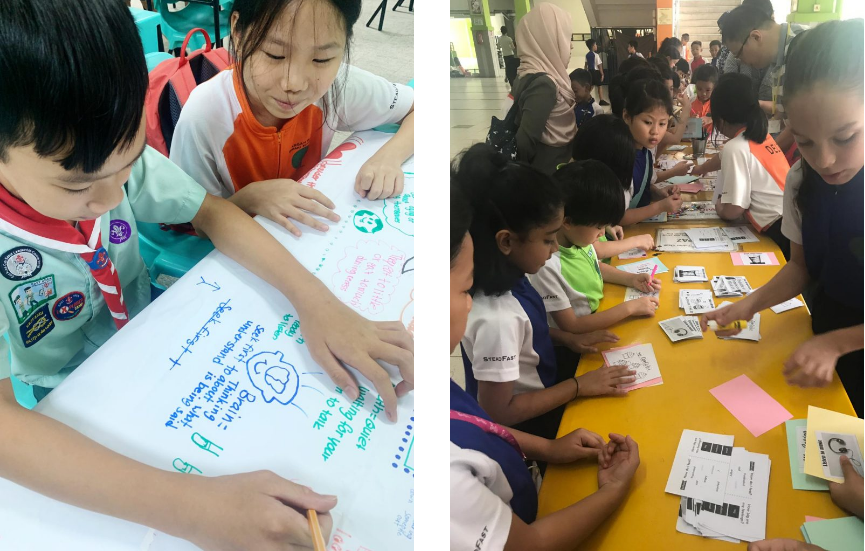 Two school children write on a large poster; separately, a group of children work on papers at a yellow table.