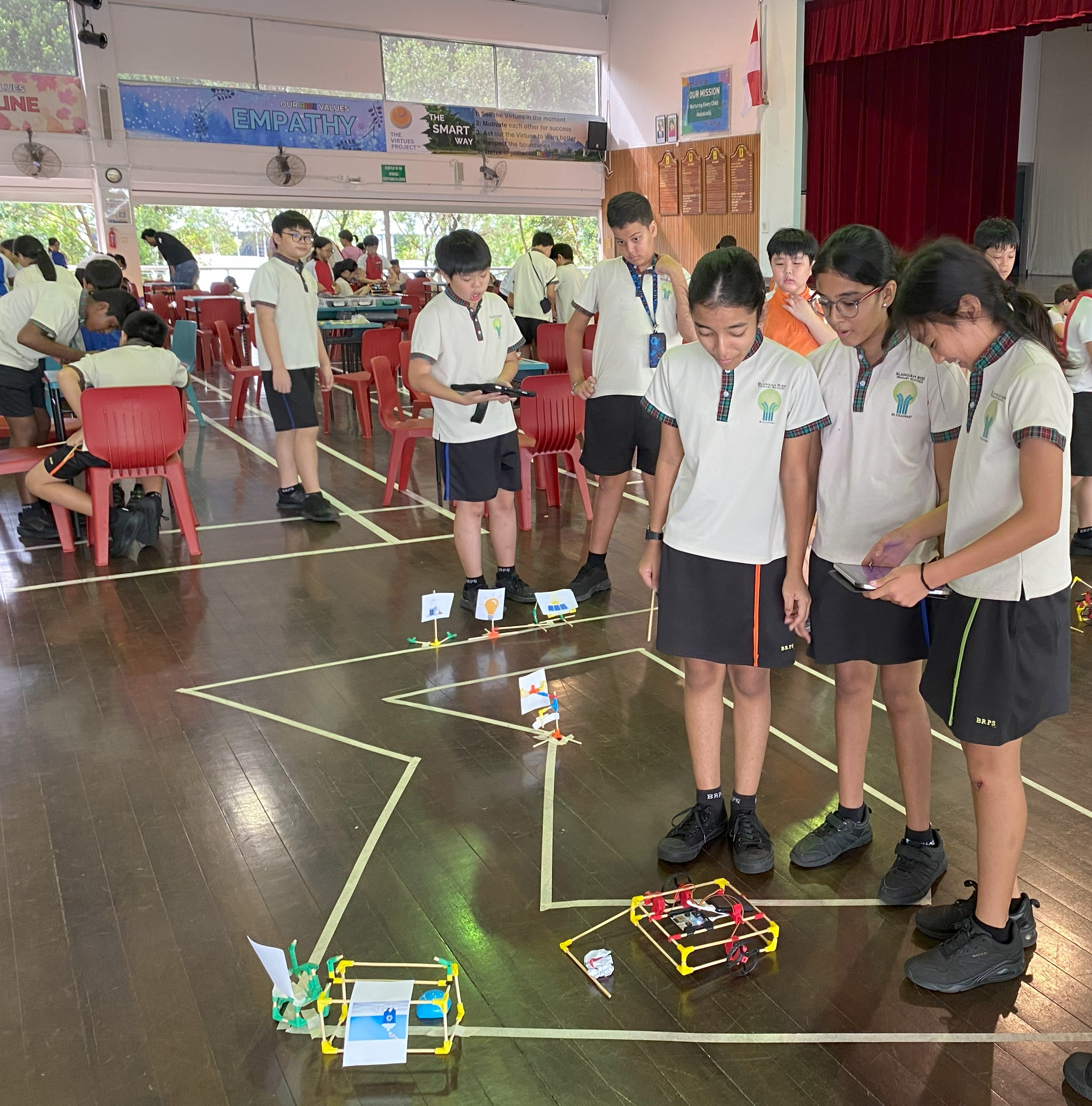 Students in school uniforms use tablets to control small robots navigating a taped track on a hall floor.