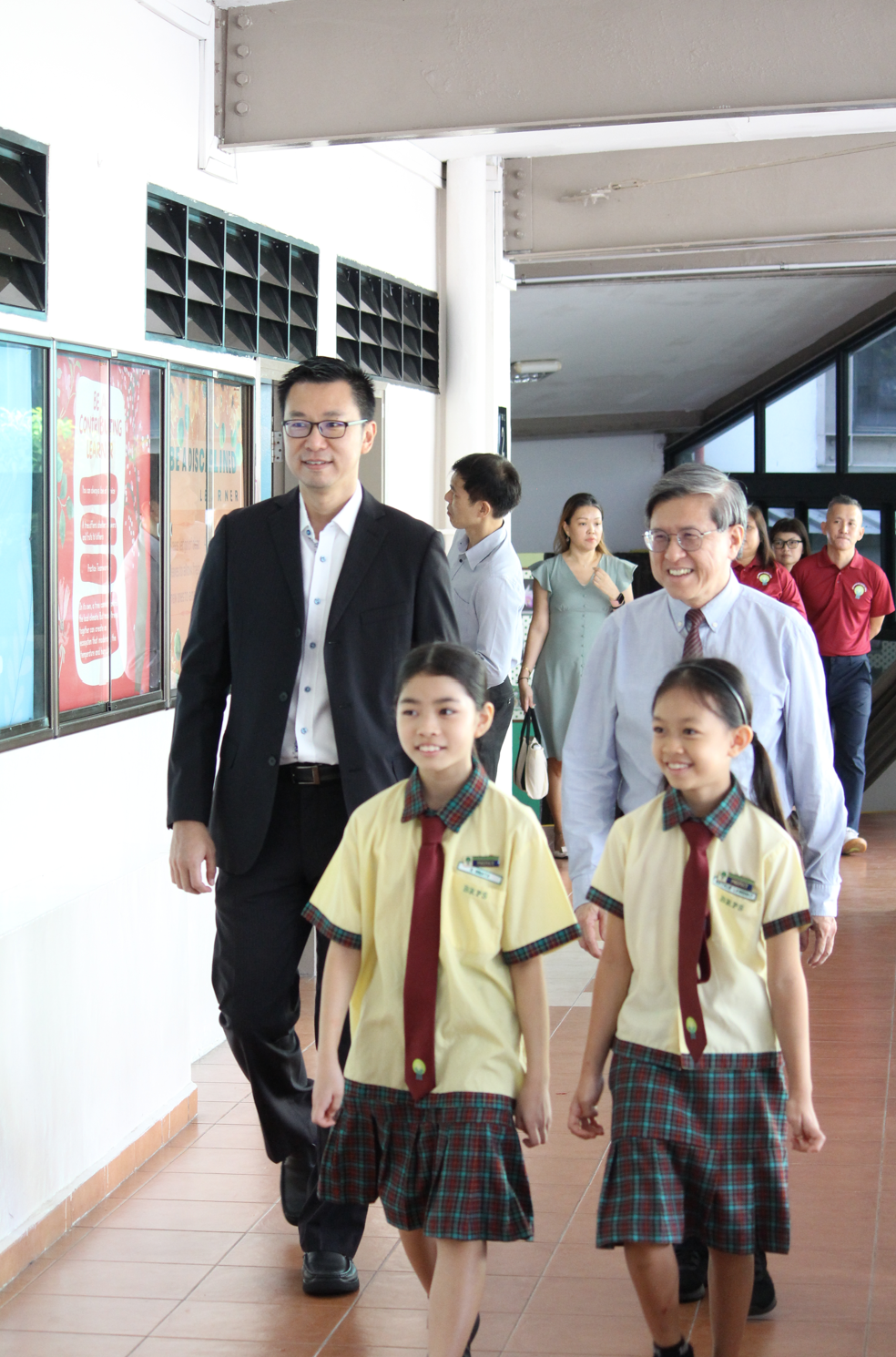 Two smiling girls in yellow plaid uniforms walk ahead of two suited men through a school corridor.