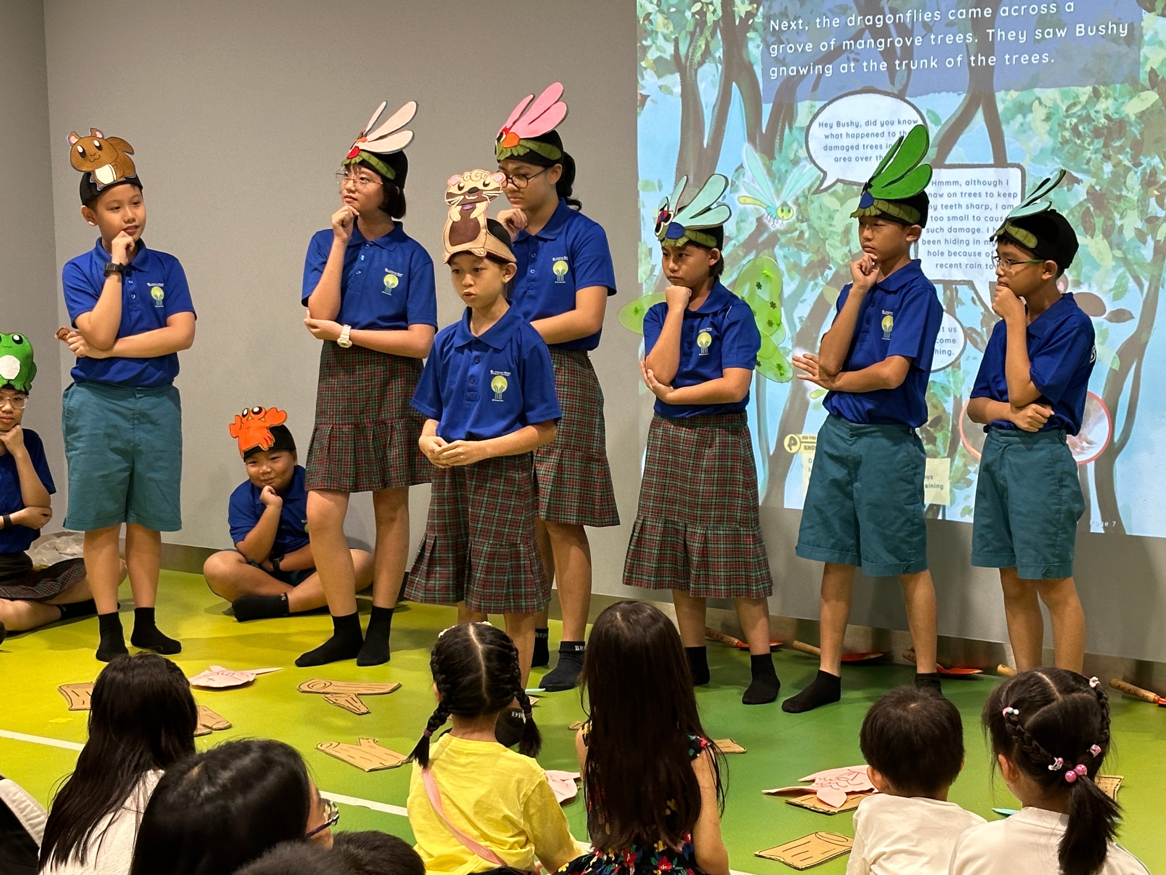 Children in blue uniforms perform a skit wearing animal hats as a forest scene is projected behind them.