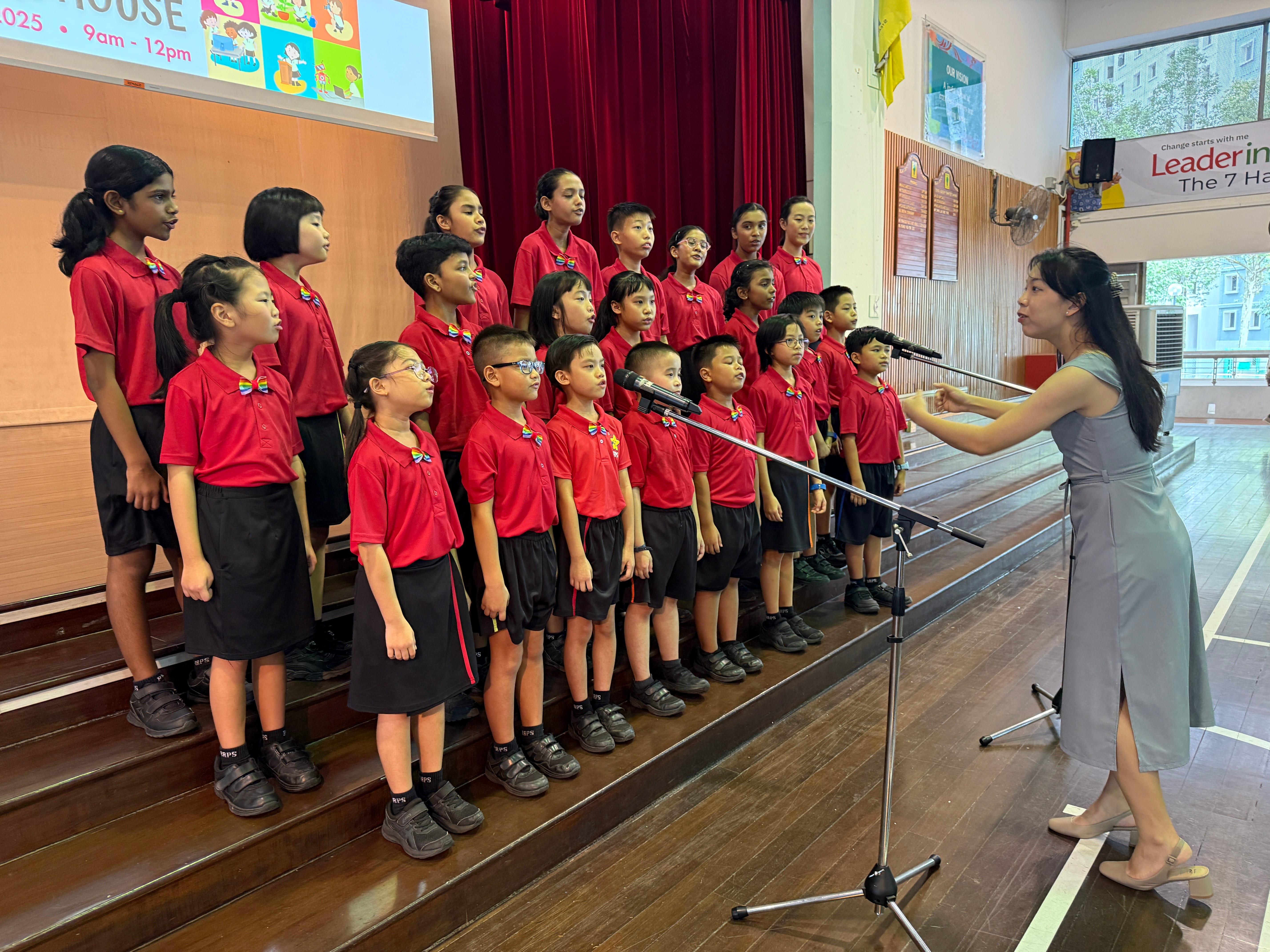 Children in red school uniforms sing on risers while a woman conducts beside two microphones.