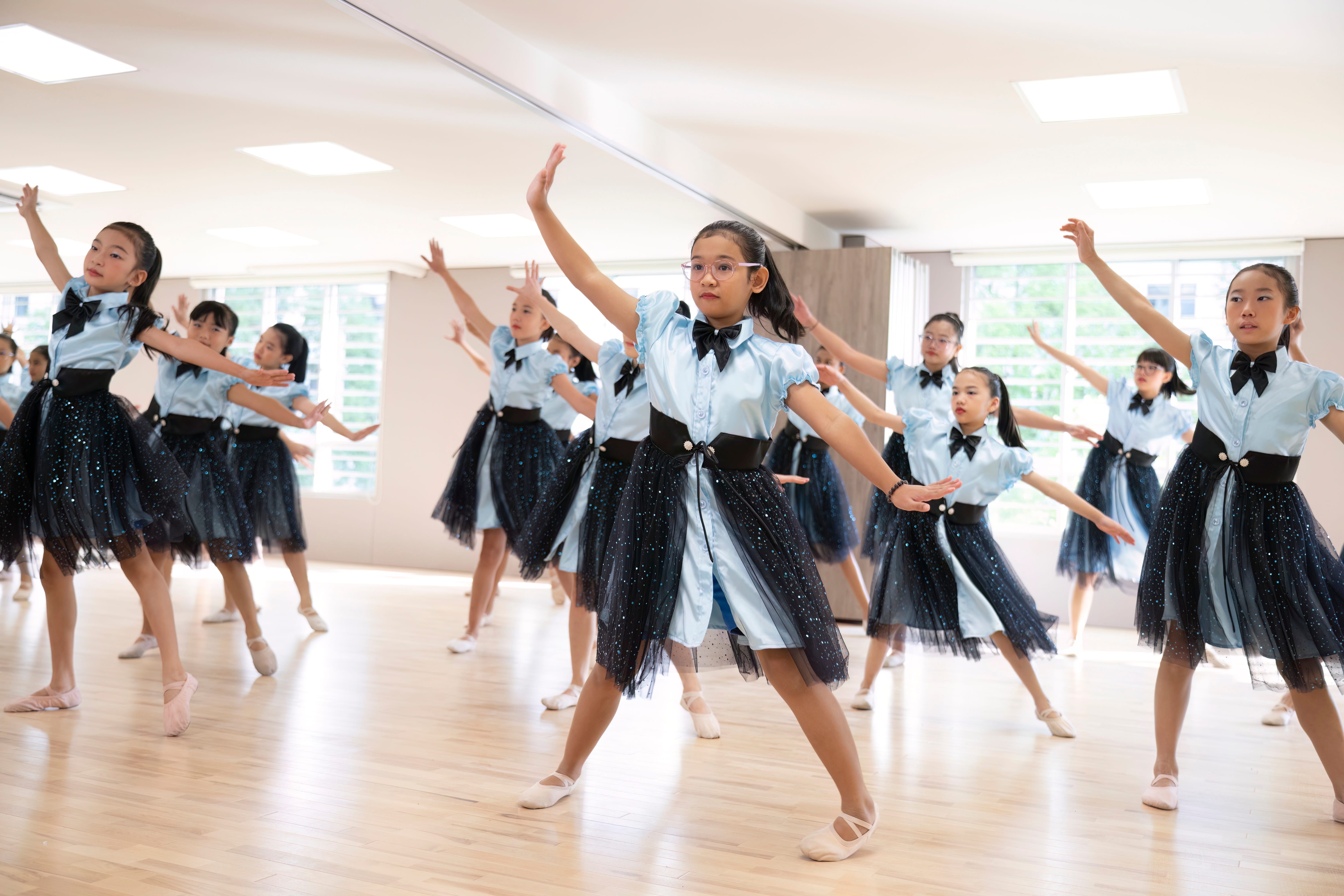 A group of young girls in matching light blue tops and dark sparkly skirts perform a synchronized dance in a studio.