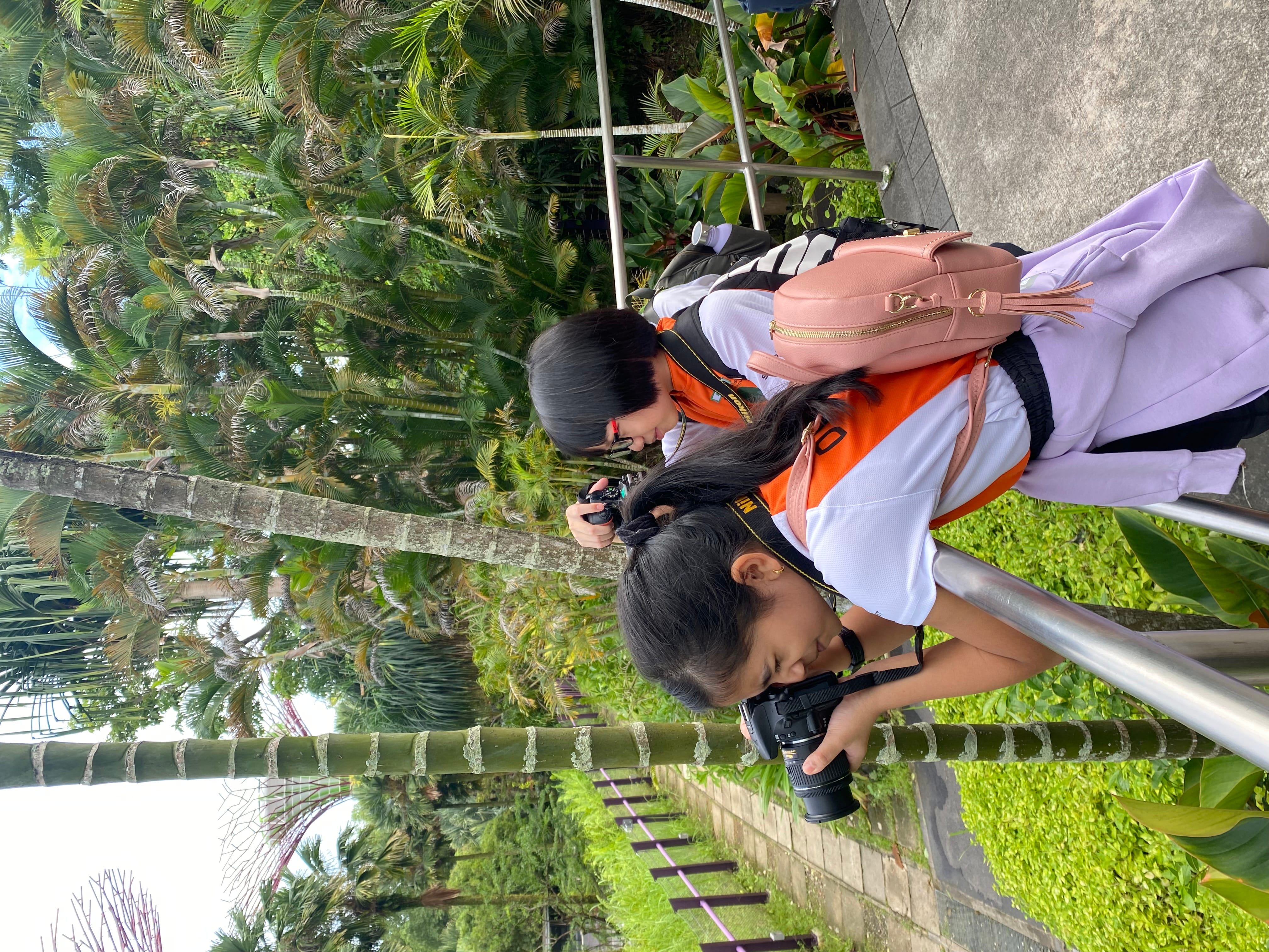 Two young girls with Nikon cameras photograph lush tropical greenery from a railing-lined path.