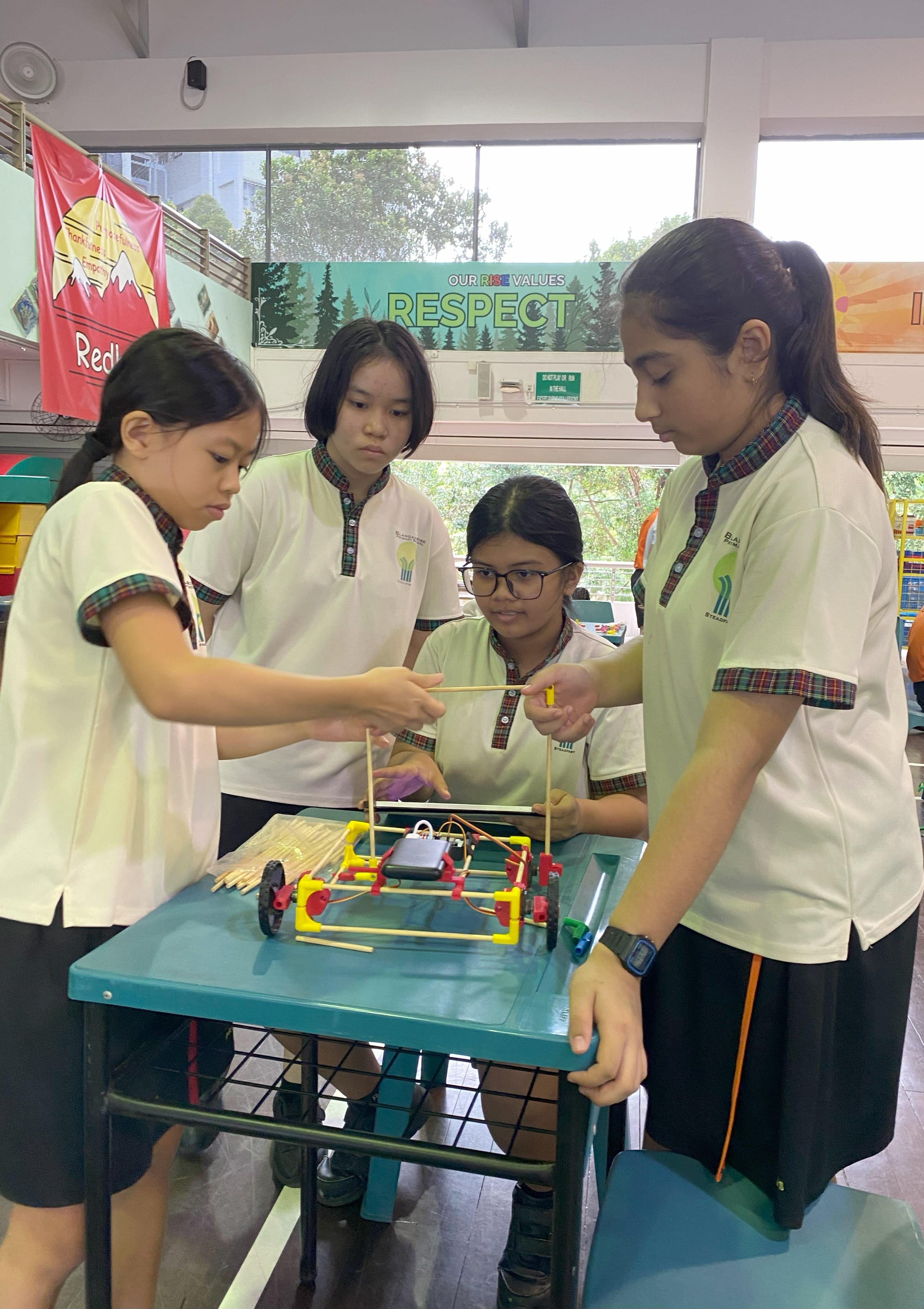 Four school girls in uniform work together assembling a small wheeled robot with wooden sticks on a teal table.