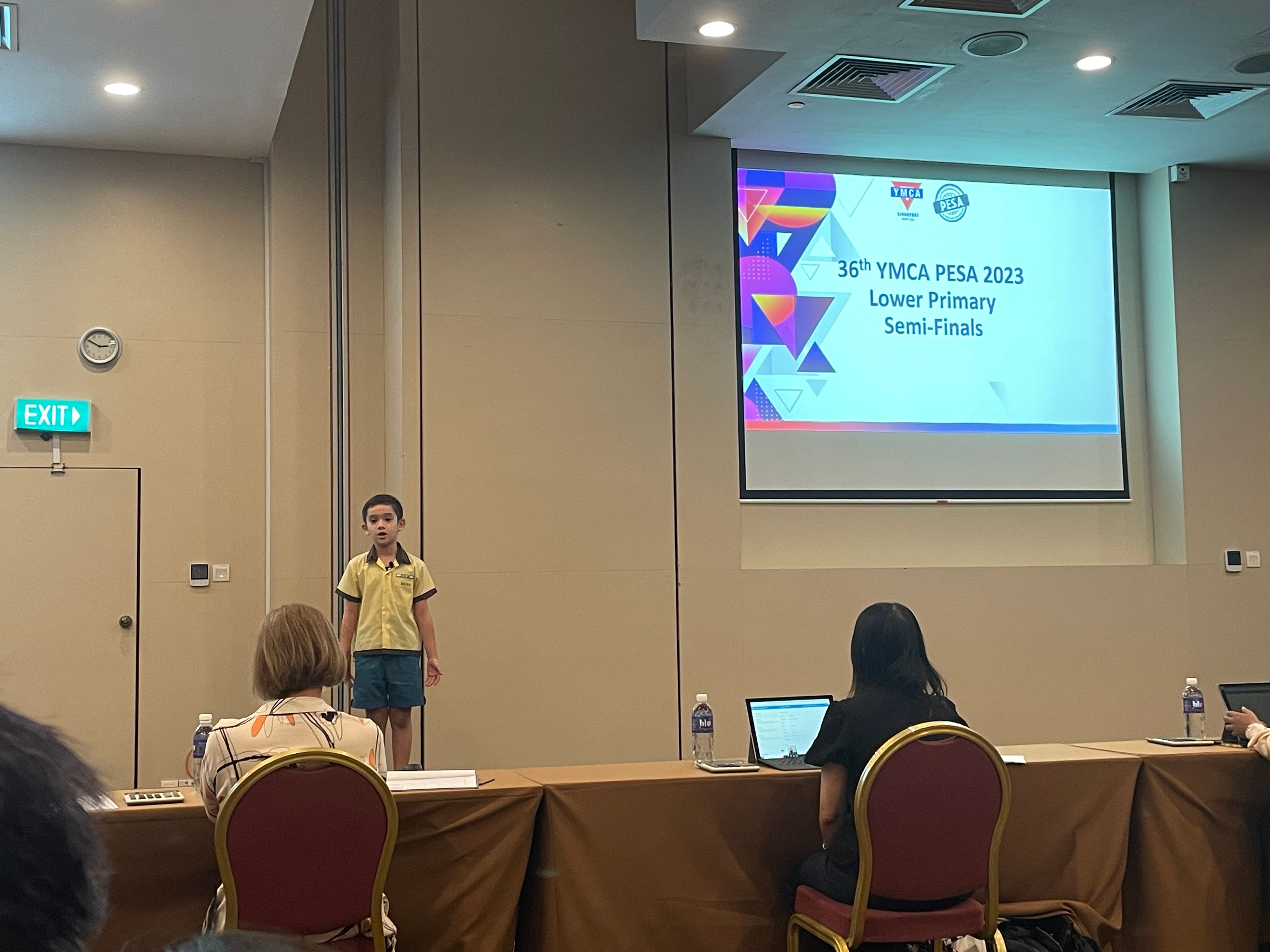 A young boy stands speaking at the 36th YMCA PESA 2023 Lower Primary Semi-Finals in a conference room.