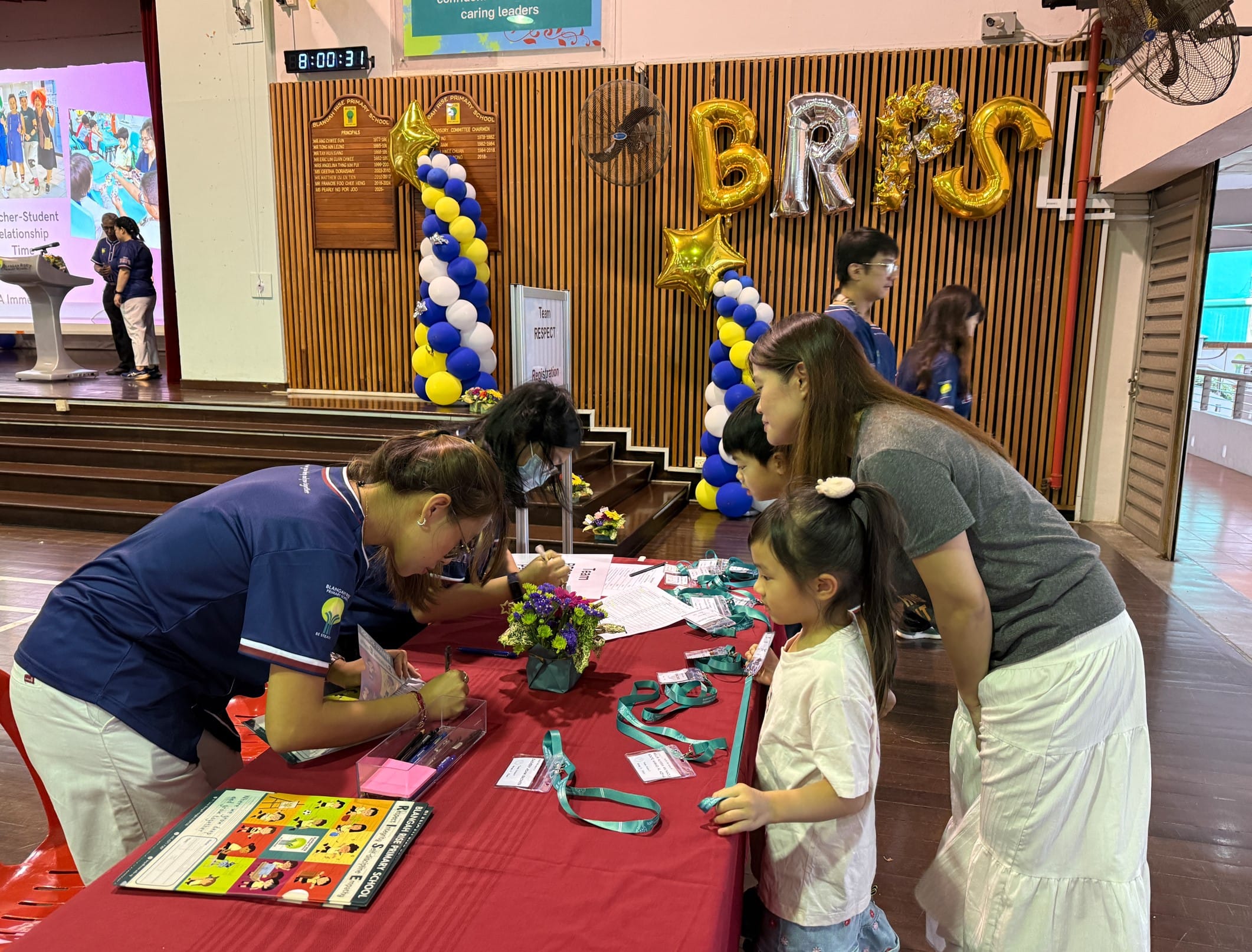 A school staff member registers a woman with two children at a red table in a decorated hall with gold and blue balloons.