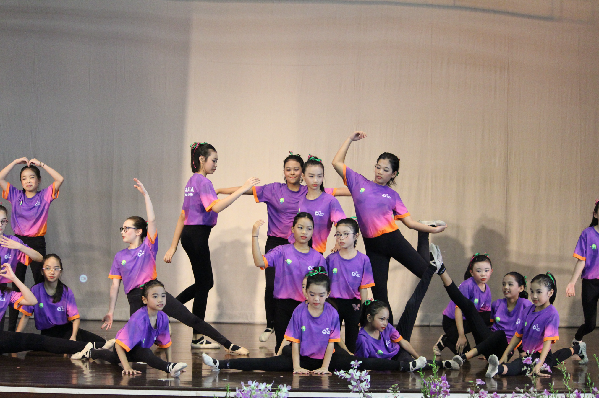 Group of girls in purple shirts and black leggings posing in dance positions on a stage.