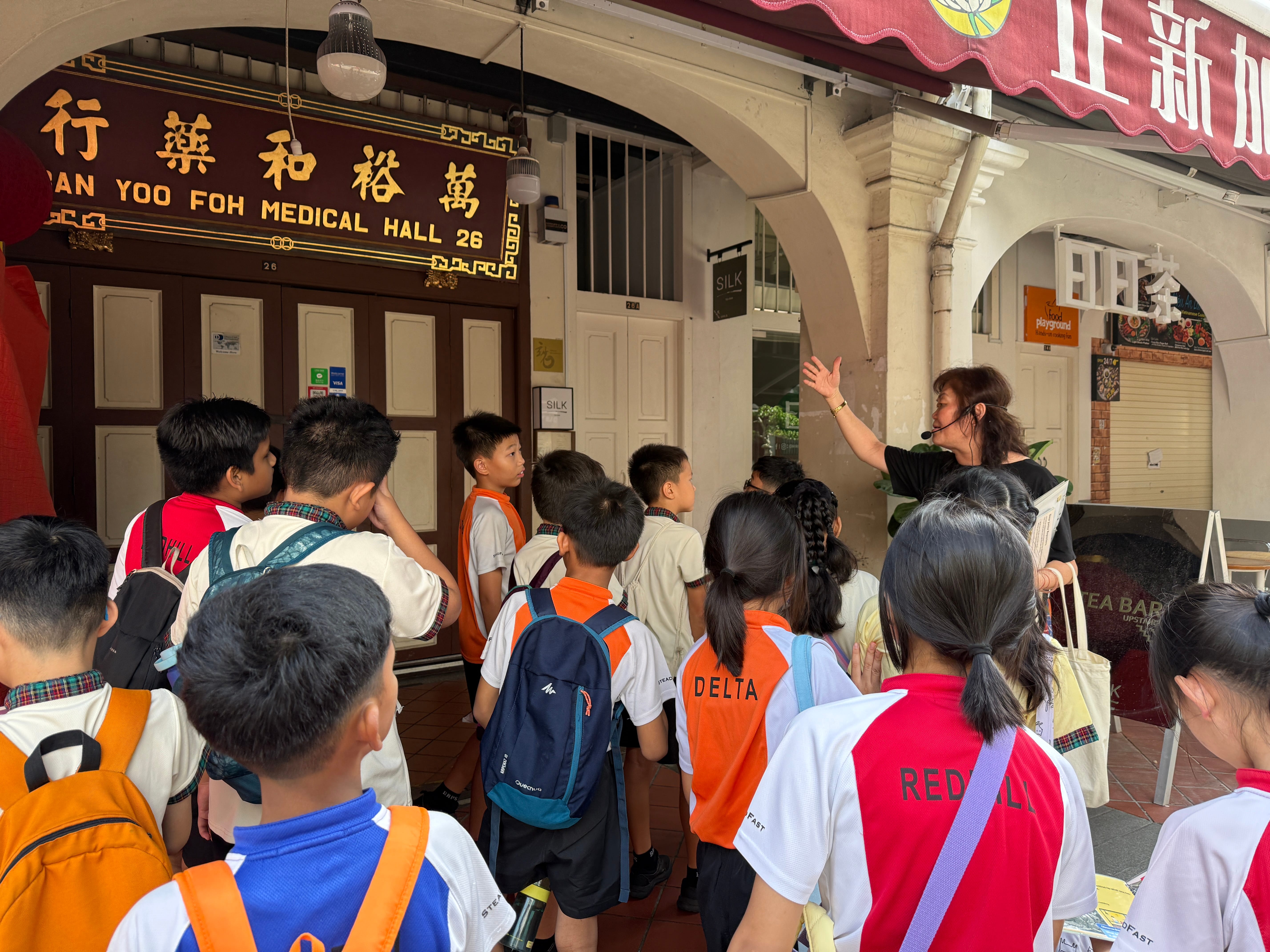 Teacher with headset gestures while students with backpacks gather under an archway beside a medical hall sign.