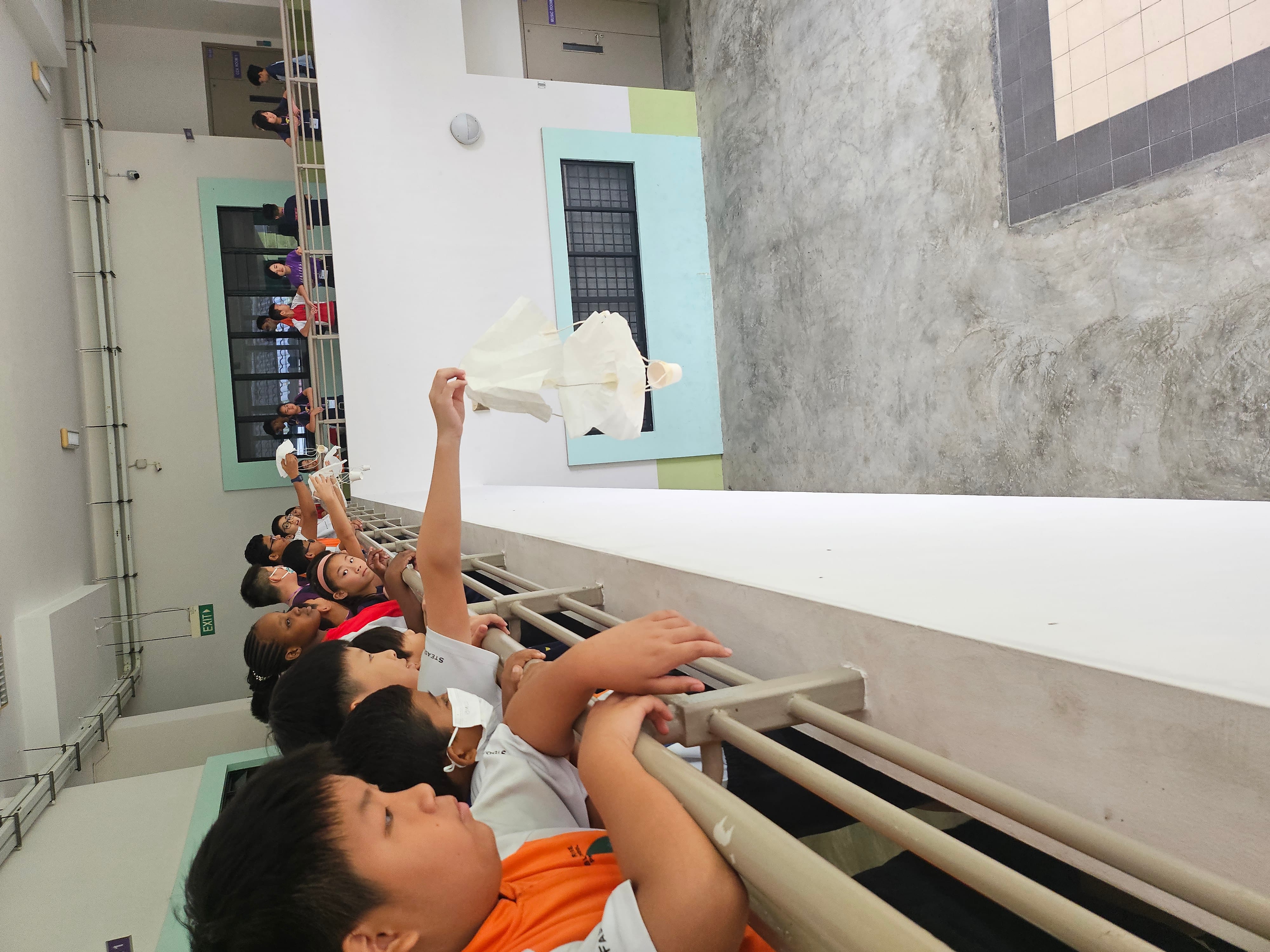 Students lean on a railing in a corridor, watching a paper parachute drop into an open courtyard.