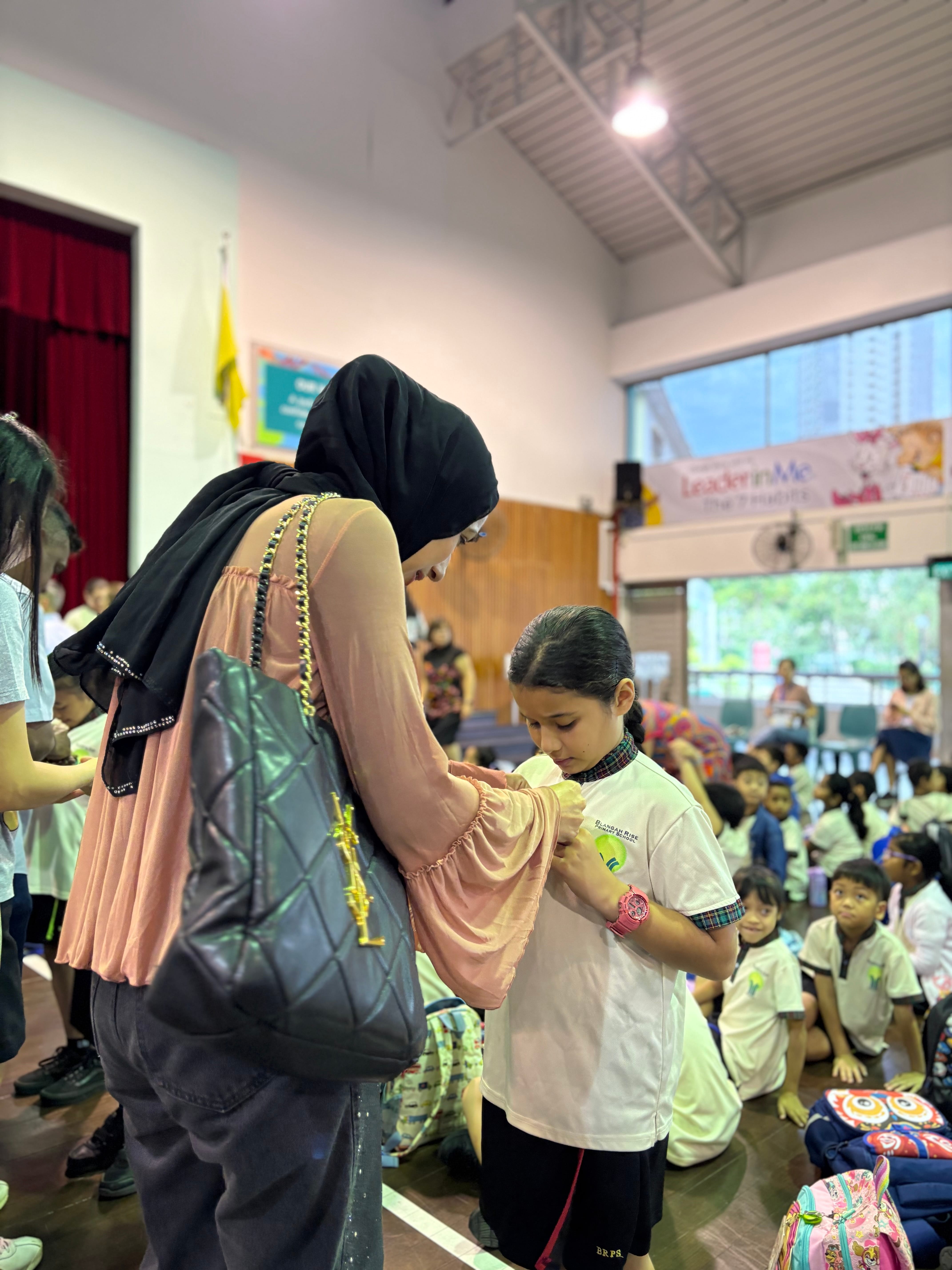 A woman in a black hijab and pink top pins something on a young girl's school uniform in a hall.