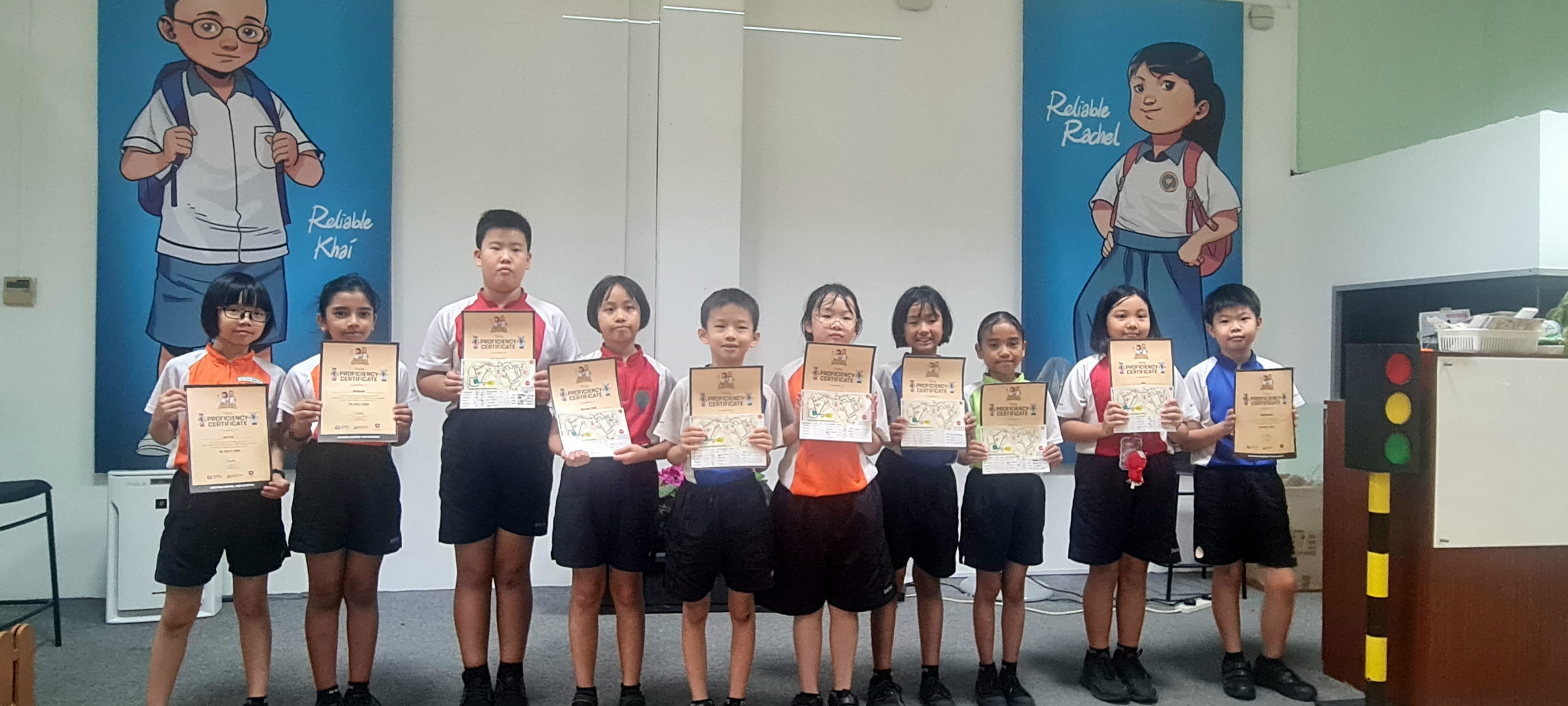Ten children stand in a row holding proficiency certificates in front of cartoon school character murals.