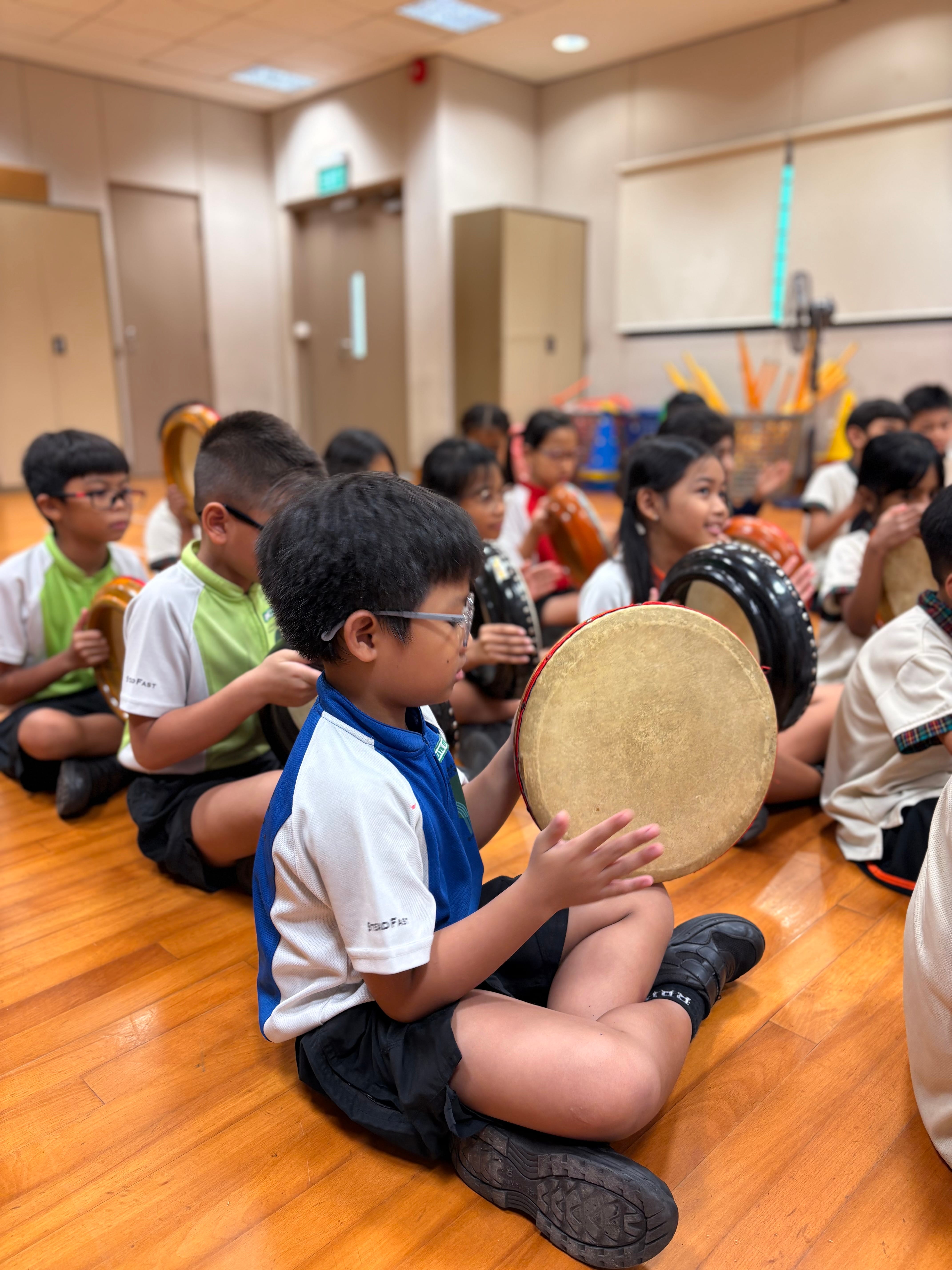 Children sit on a wooden floor in a classroom, each holding a round hand drum during a music lesson.