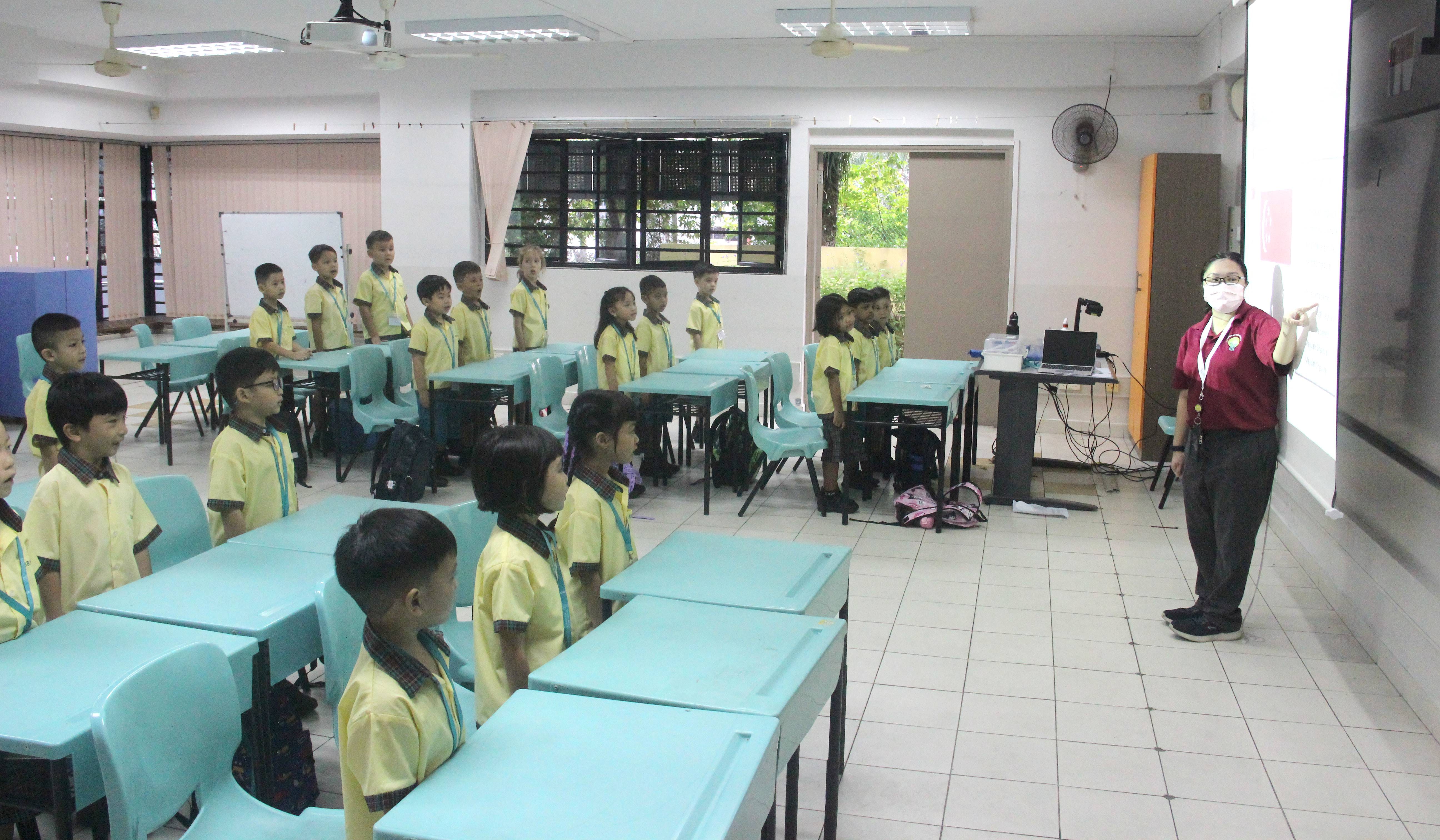 A masked teacher in red points at a screen while young students in yellow uniforms stand at their teal desks.