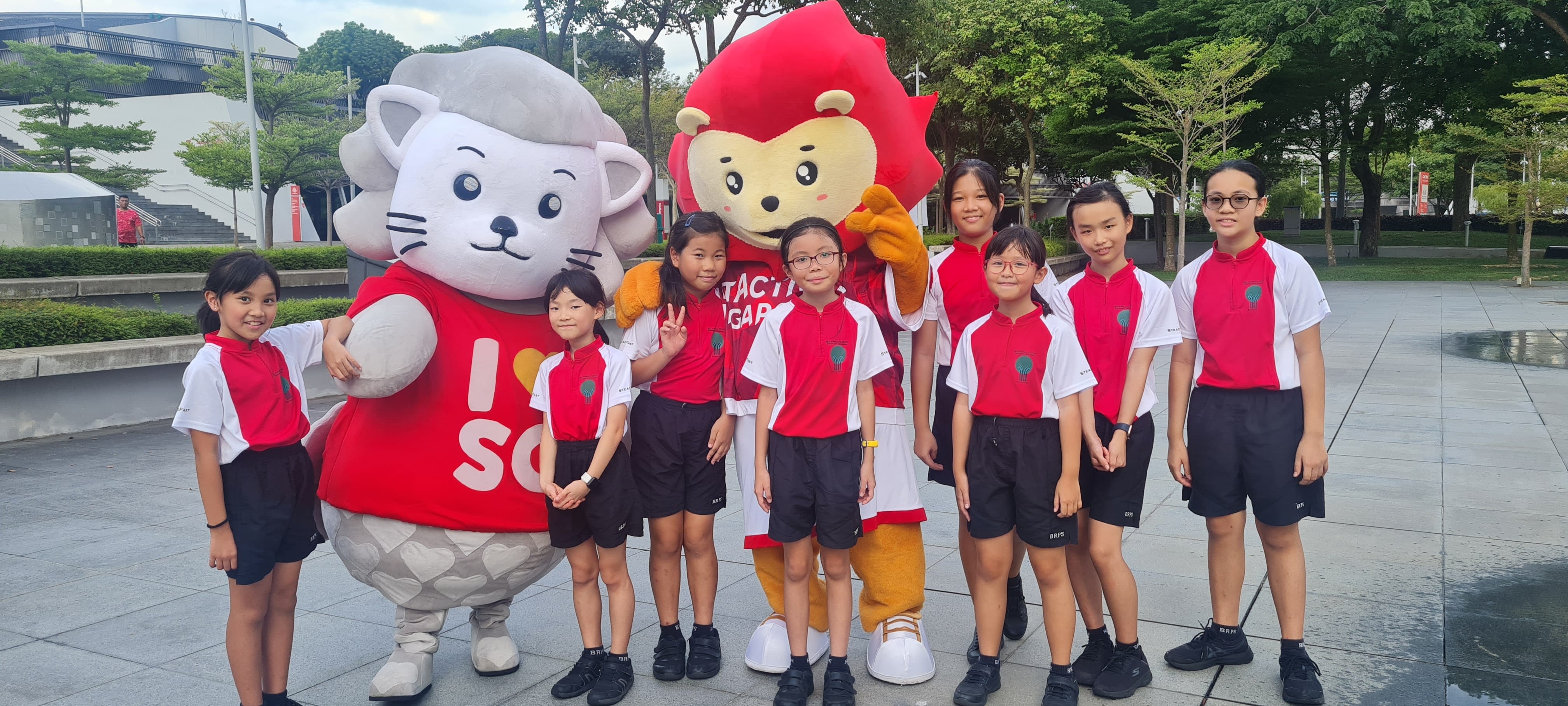 Eight students in red-and-white uniforms pose with a Merlion and lion mascot in a park plaza.