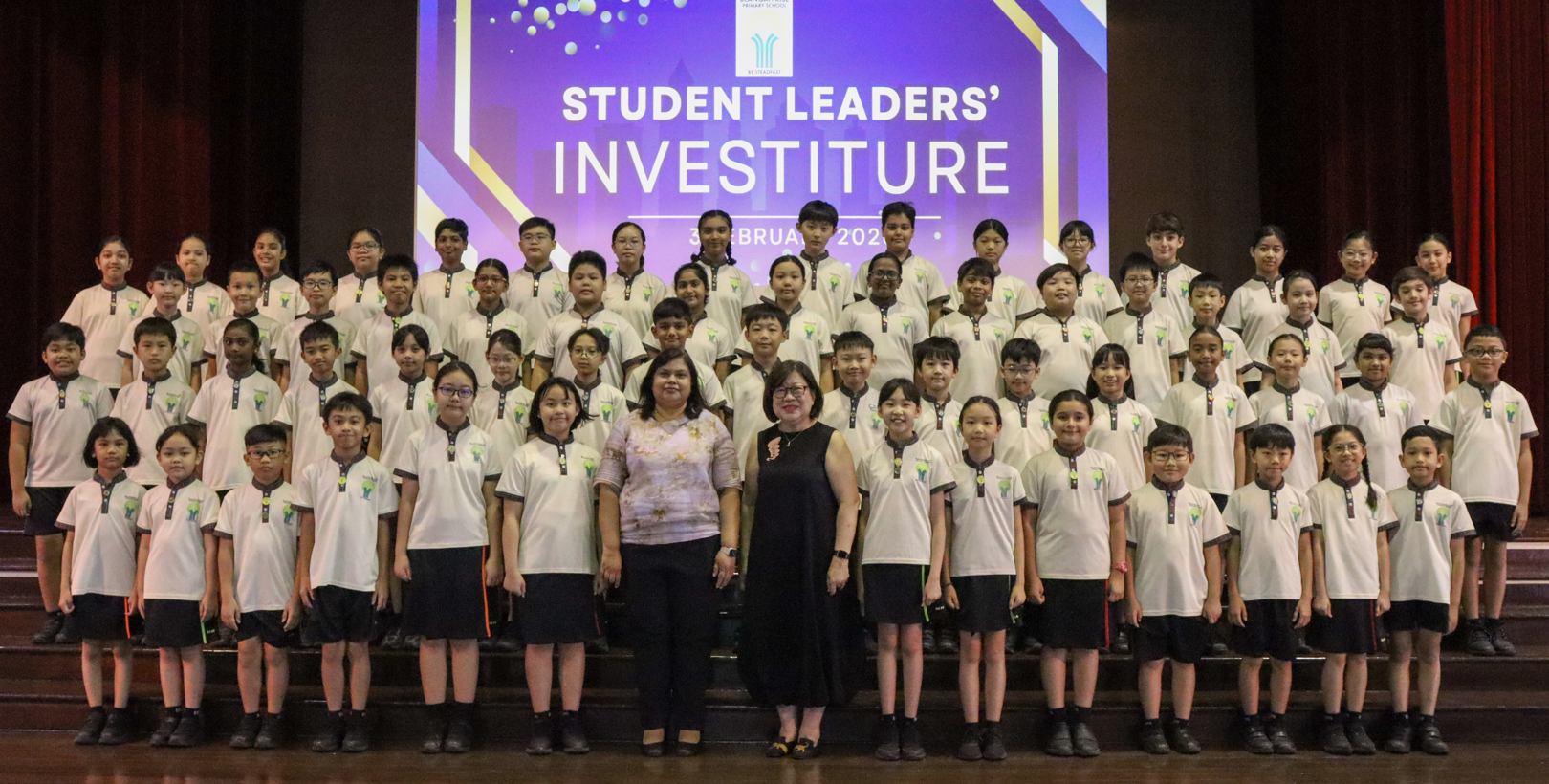 Large group of children in white uniforms pose with two adults at a Student Leaders Investiture ceremony on stage.