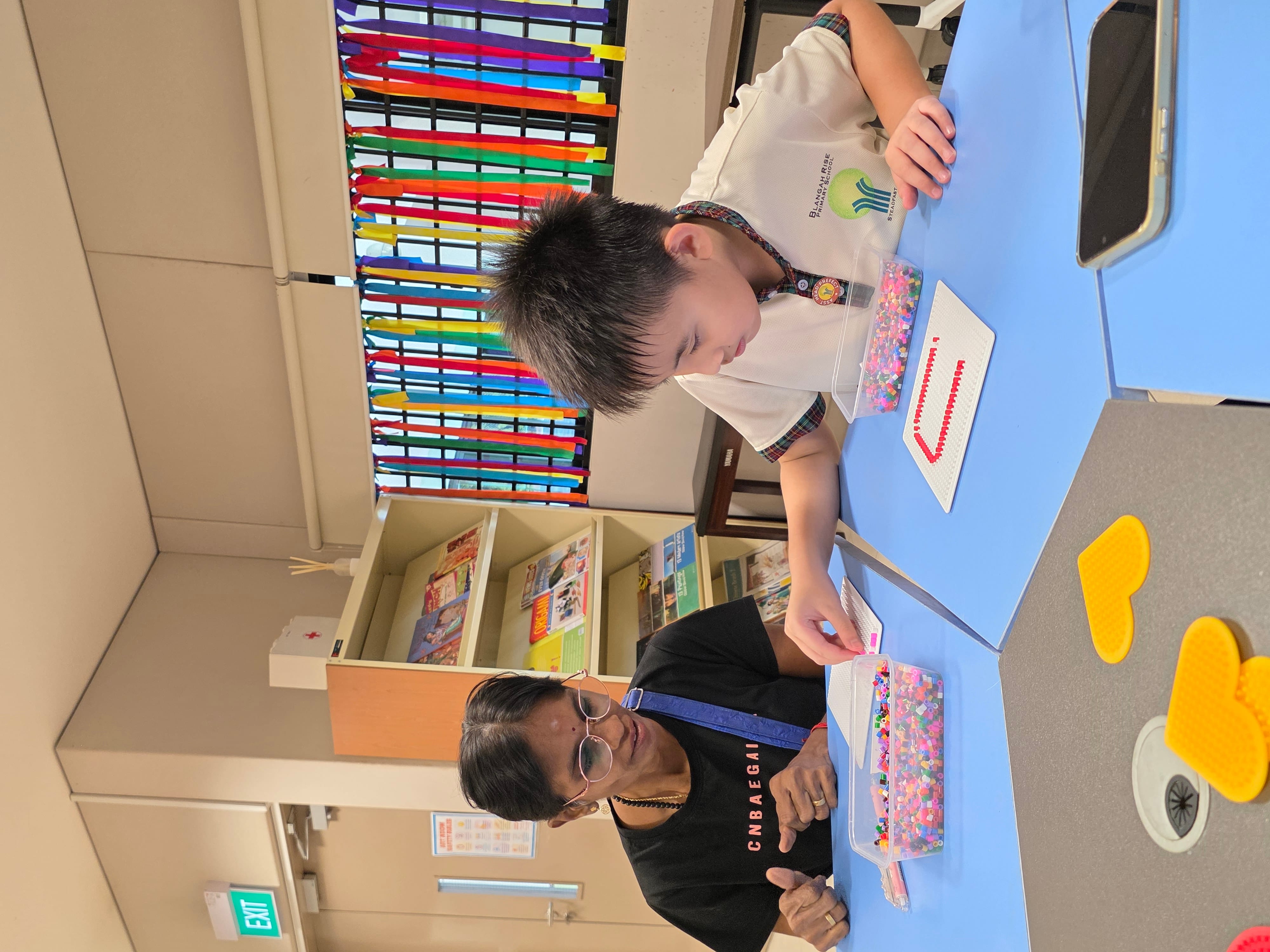 A woman and a young boy sit at a blue table working with colorful beads on pegboards in a classroom.