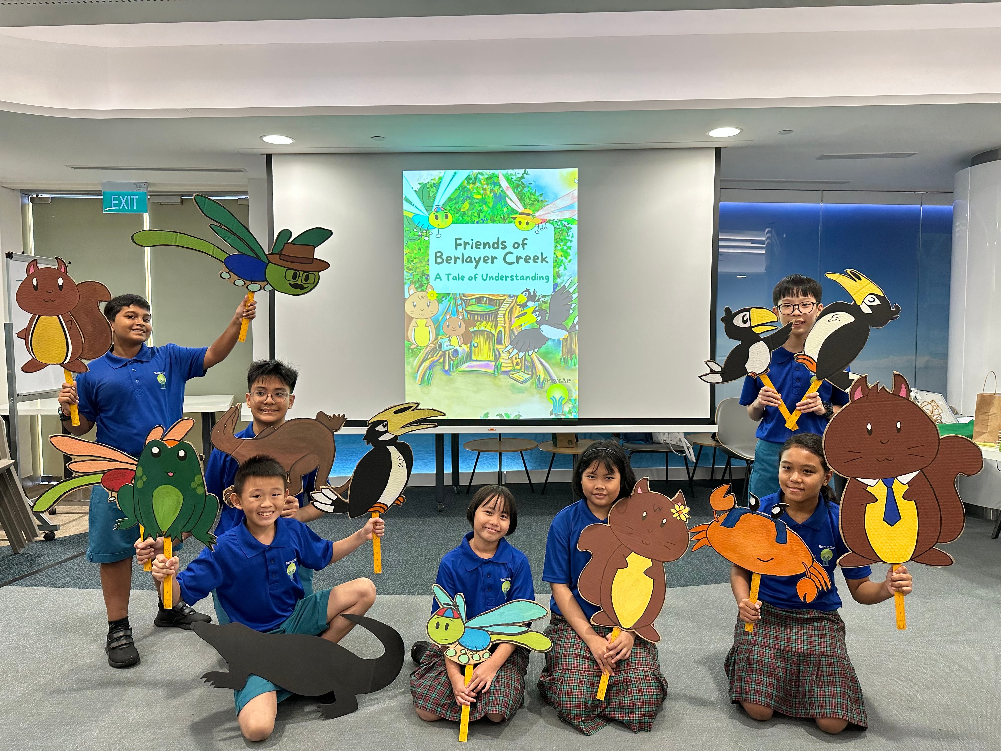 Children in blue uniforms pose with animal cutouts before a screen showing a storybook cover.