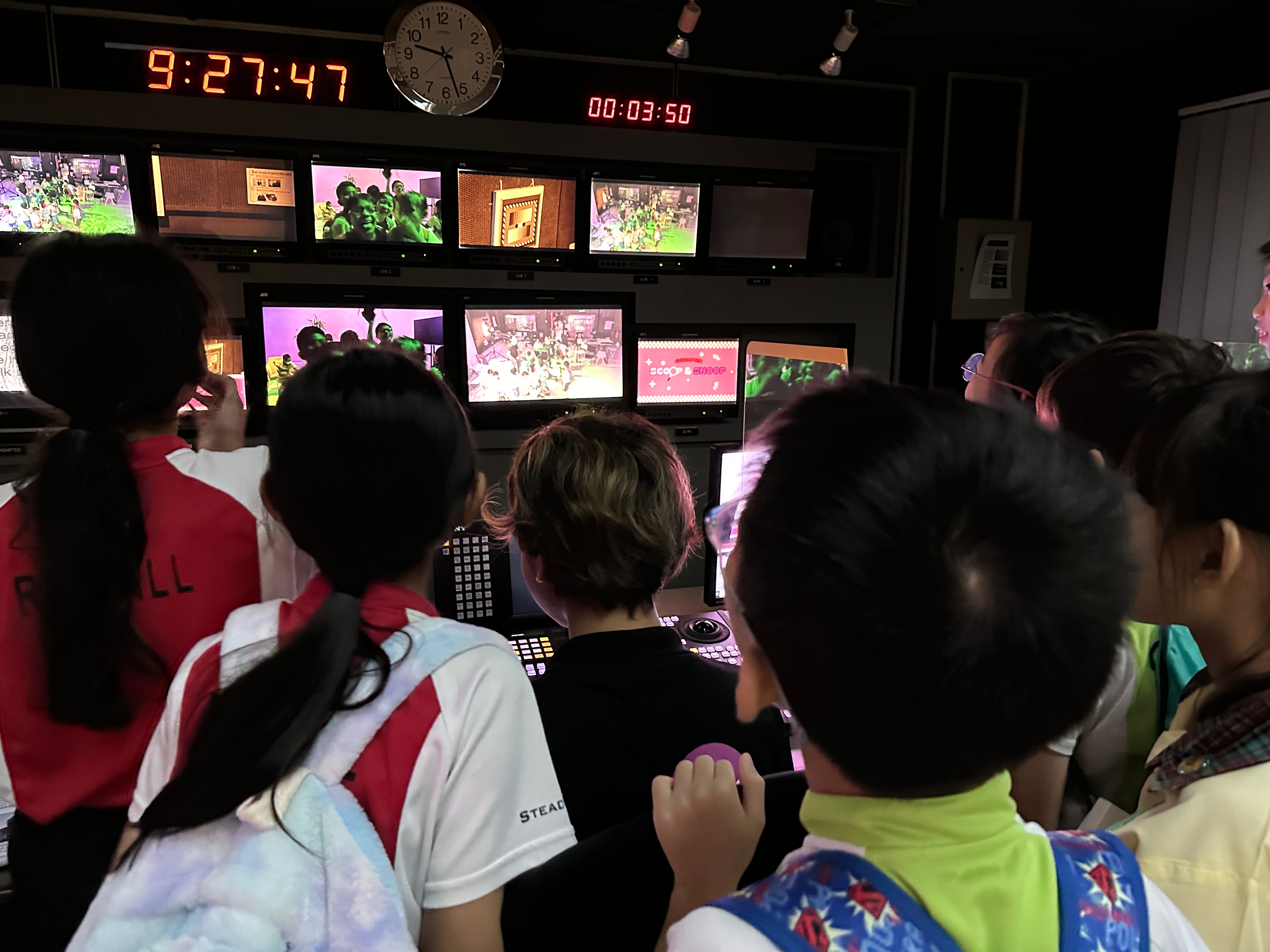 Children stand in a dark TV control room watching multiple monitors showing live feeds, with clocks displaying 9:27:47.