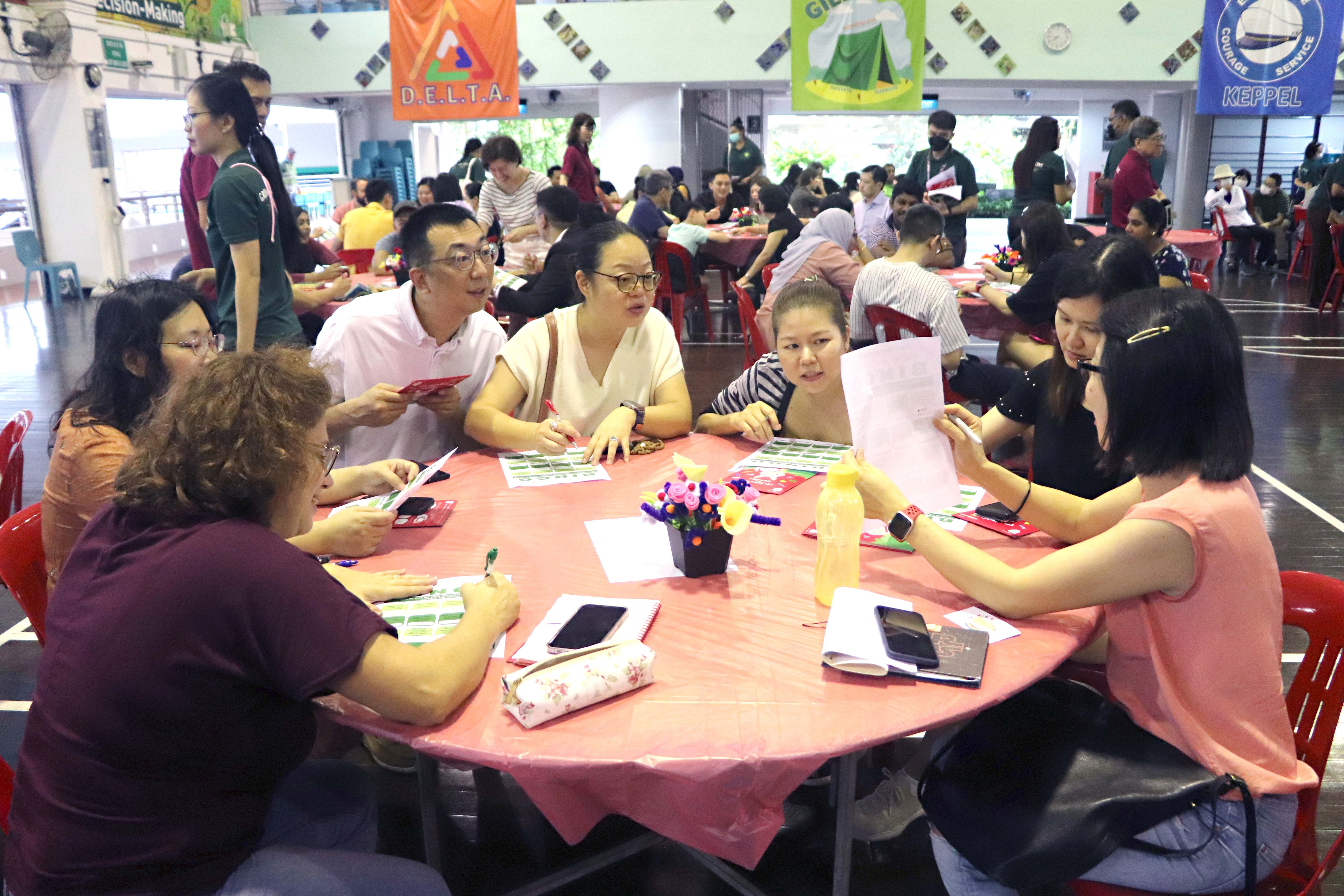 Several adults sit around a pink-covered round table in a busy hall, discussing papers and cards.