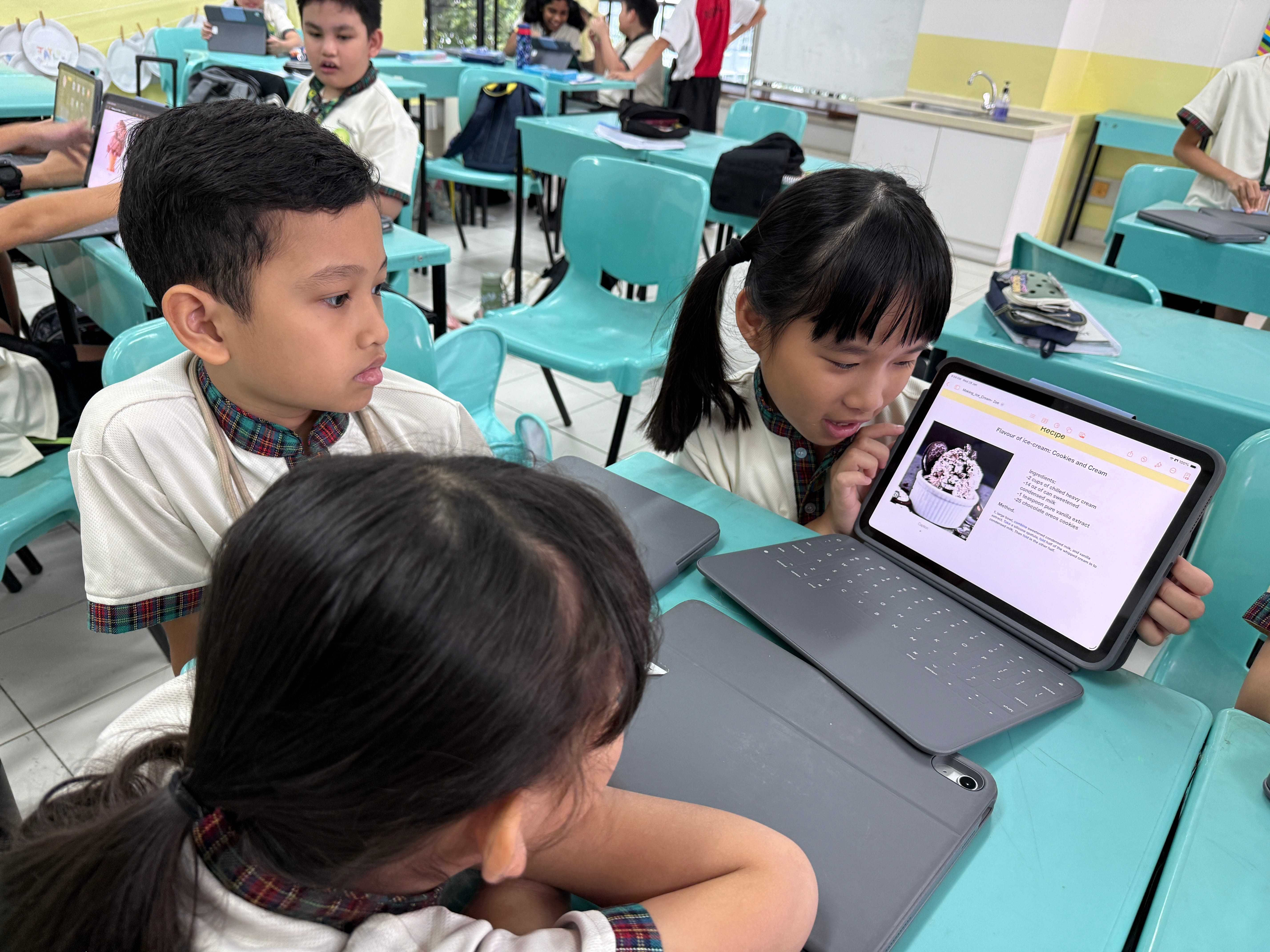Three schoolchildren in uniforms sit at desks, looking at a tablet showing a dessert recipe page.