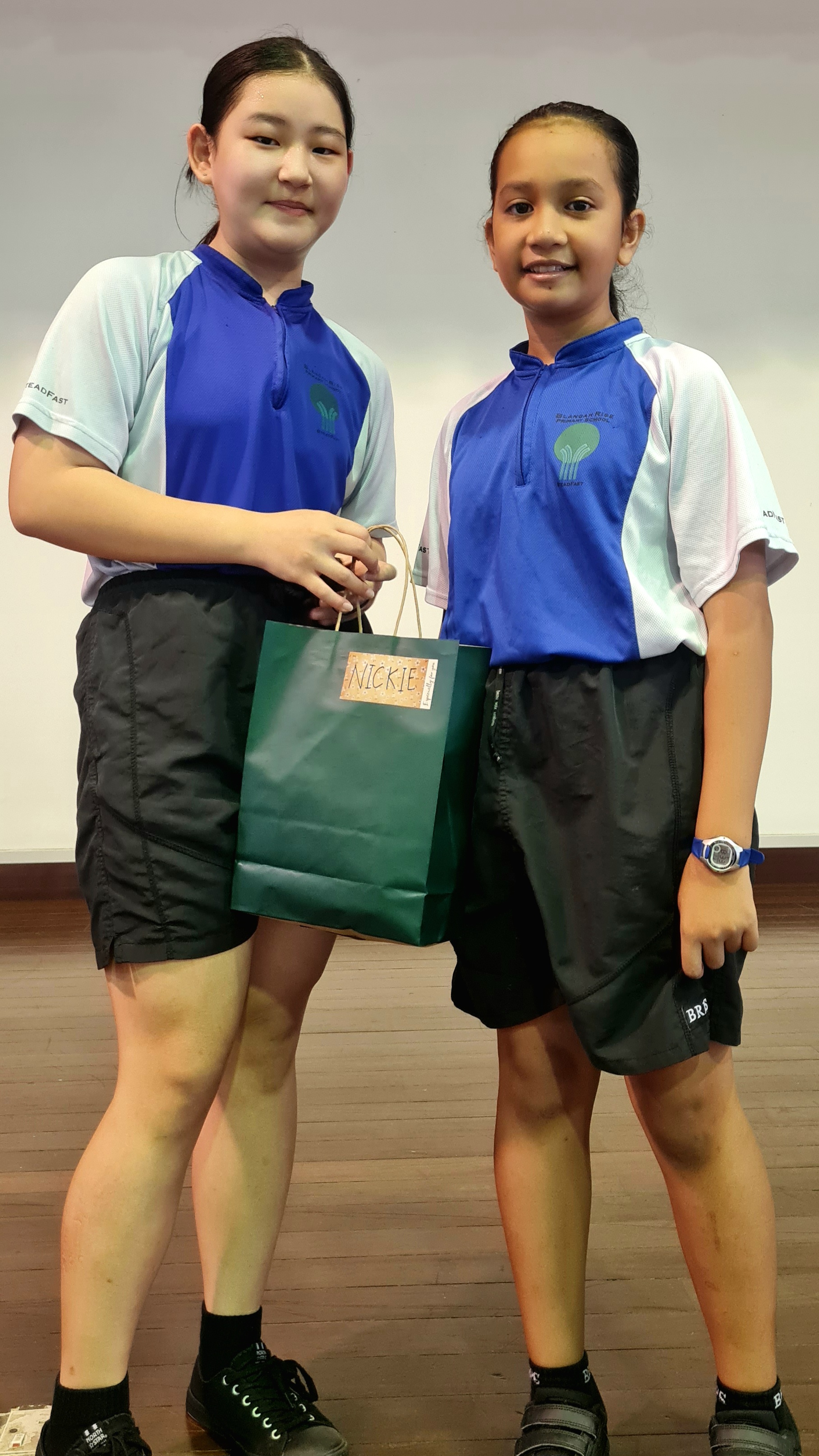 Two girls in blue and white school uniforms stand together holding a green gift bag labeled "Nickie."