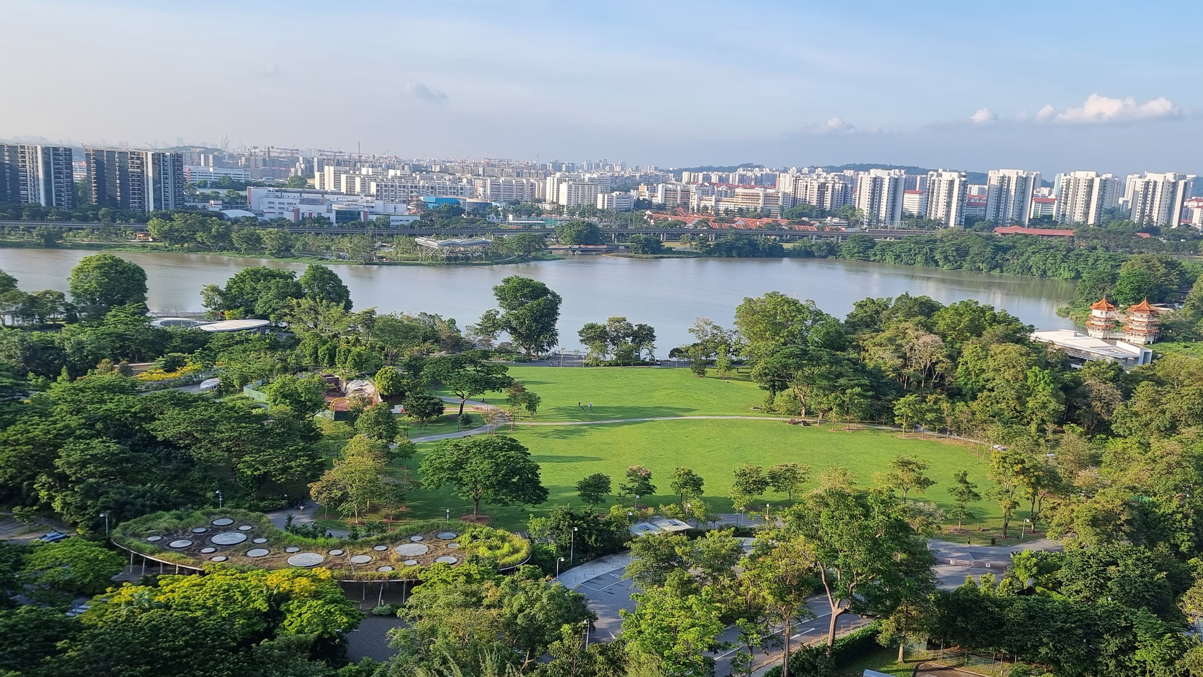 Aerial view of a large green park with a lake, dense trees, and a city skyline of white buildings in the background.