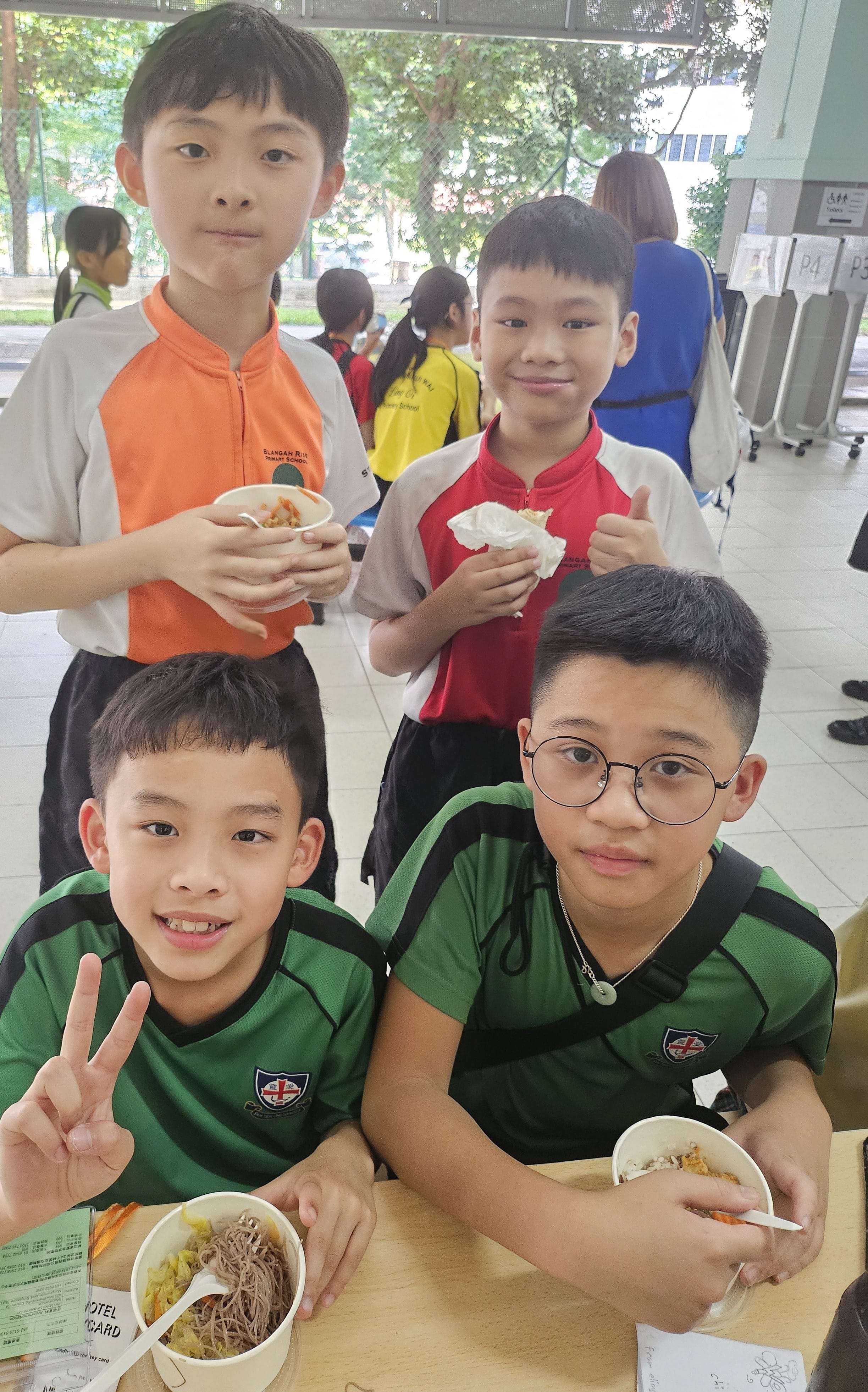 Four young boys in school uniforms eating noodle bowls at a canteen table, one flashing a peace sign.