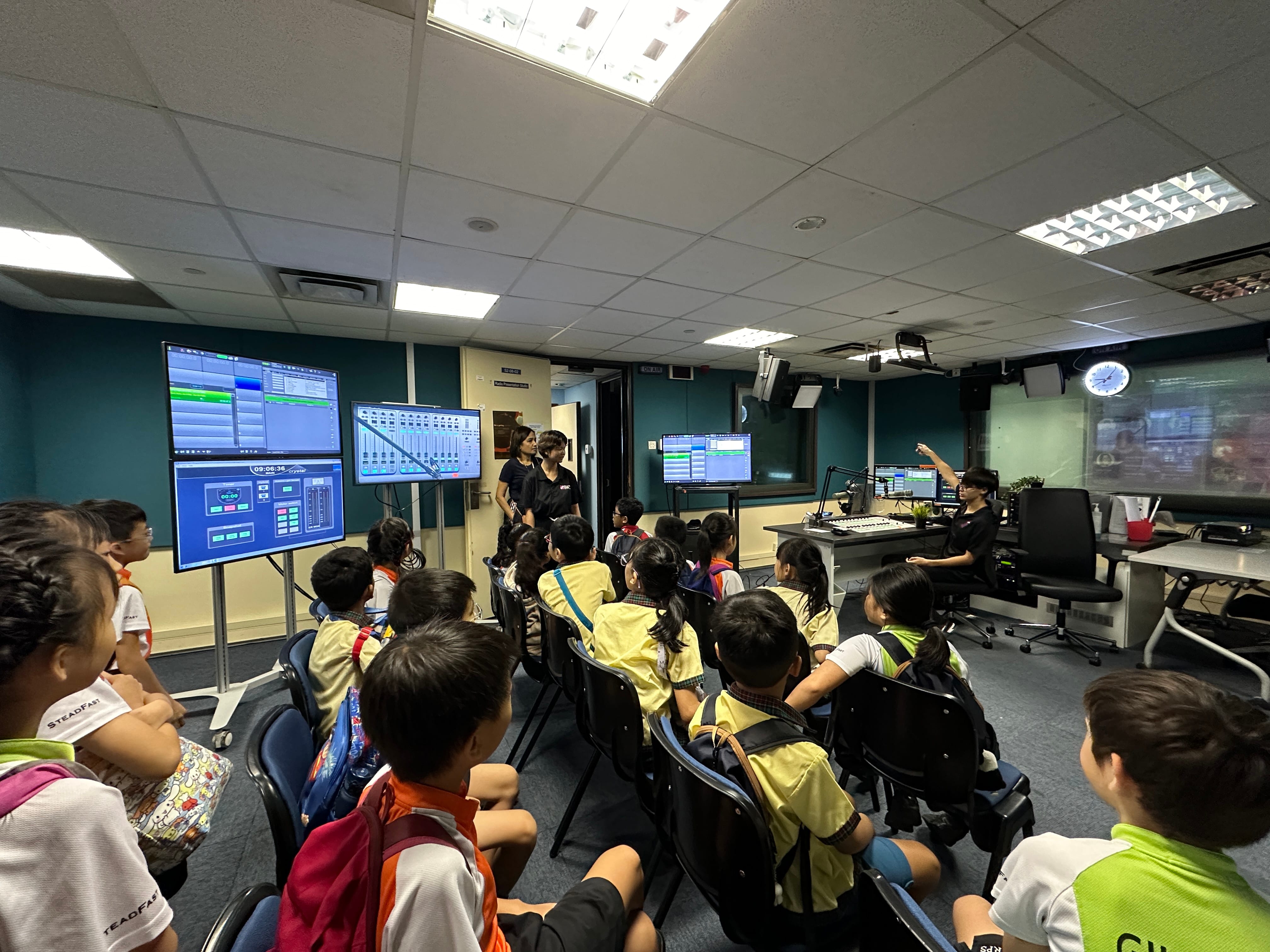 Children in yellow uniforms sit in chairs watching a presenter in a broadcast control room with multiple screens.