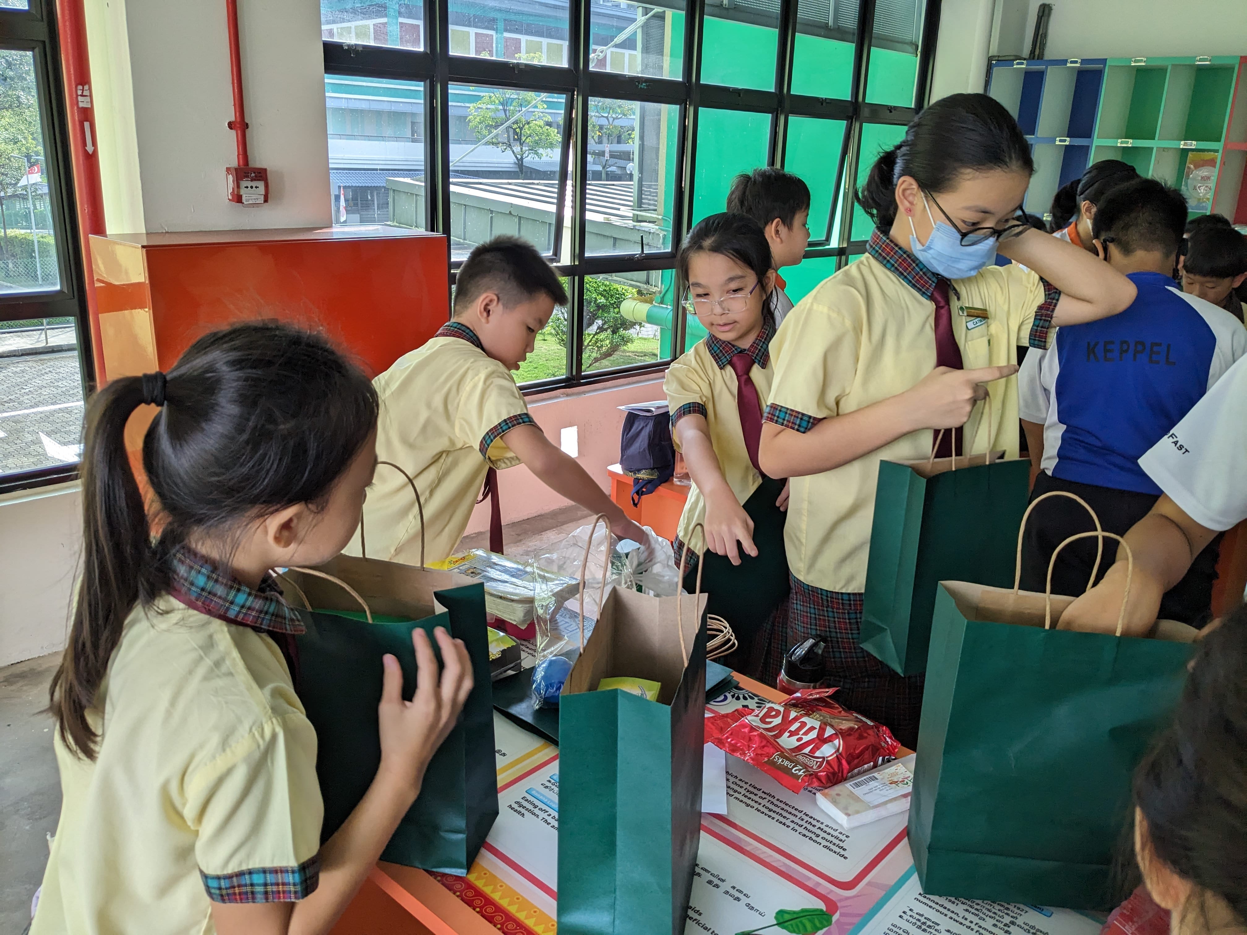 Several children in matching school uniforms sort items into green and brown paper bags on a table indoors.