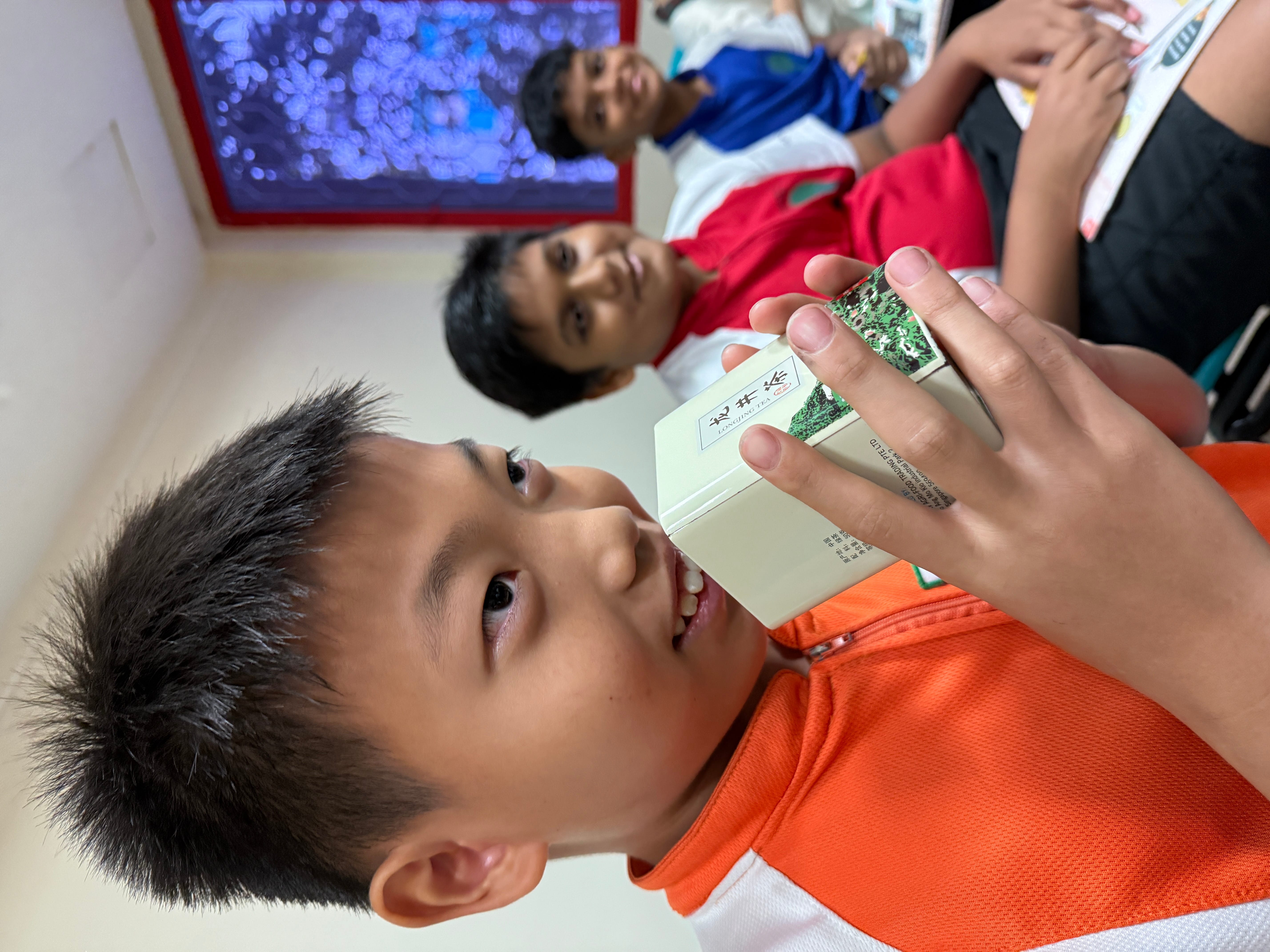 A boy in an orange shirt holds a small Chinese tea box, smiling, with two other boys seated behind him in a classroom.