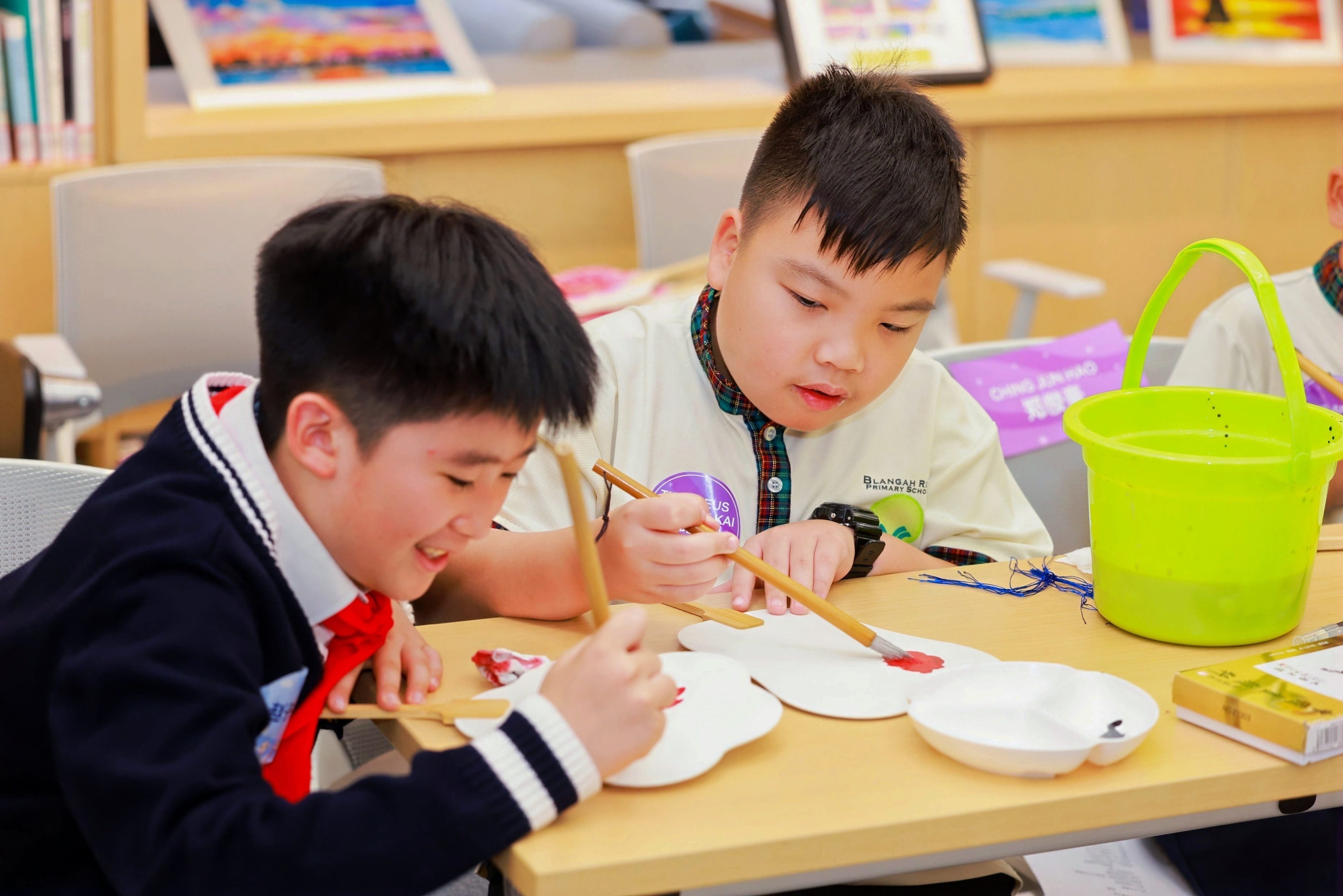 Two young boys in school uniforms paint on white paper shapes at a desk with brushes and a green bucket nearby.