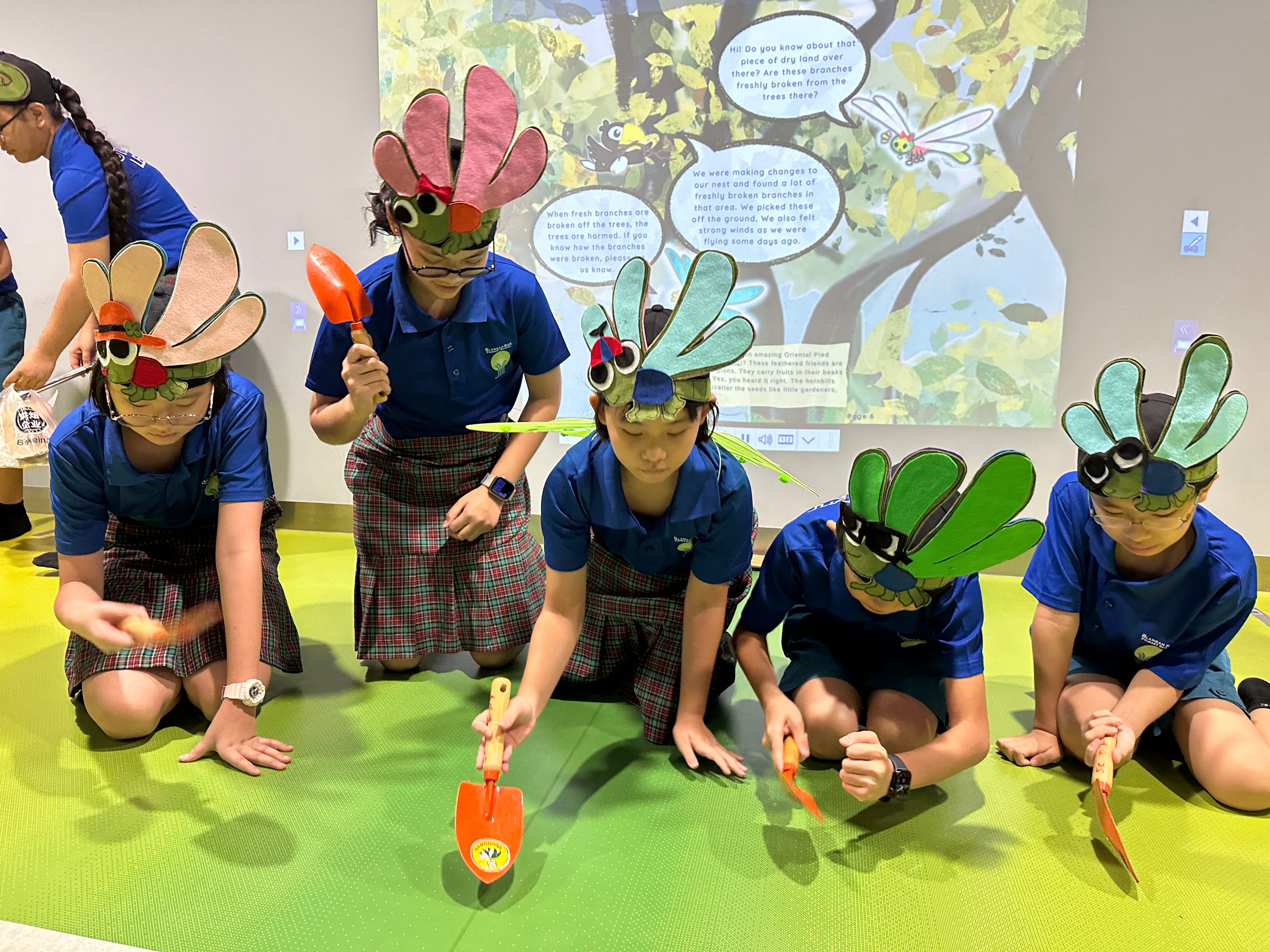 Four children in blue uniforms wear colorful bird masks and hold small shovels, kneeling before a projected storybook screen.