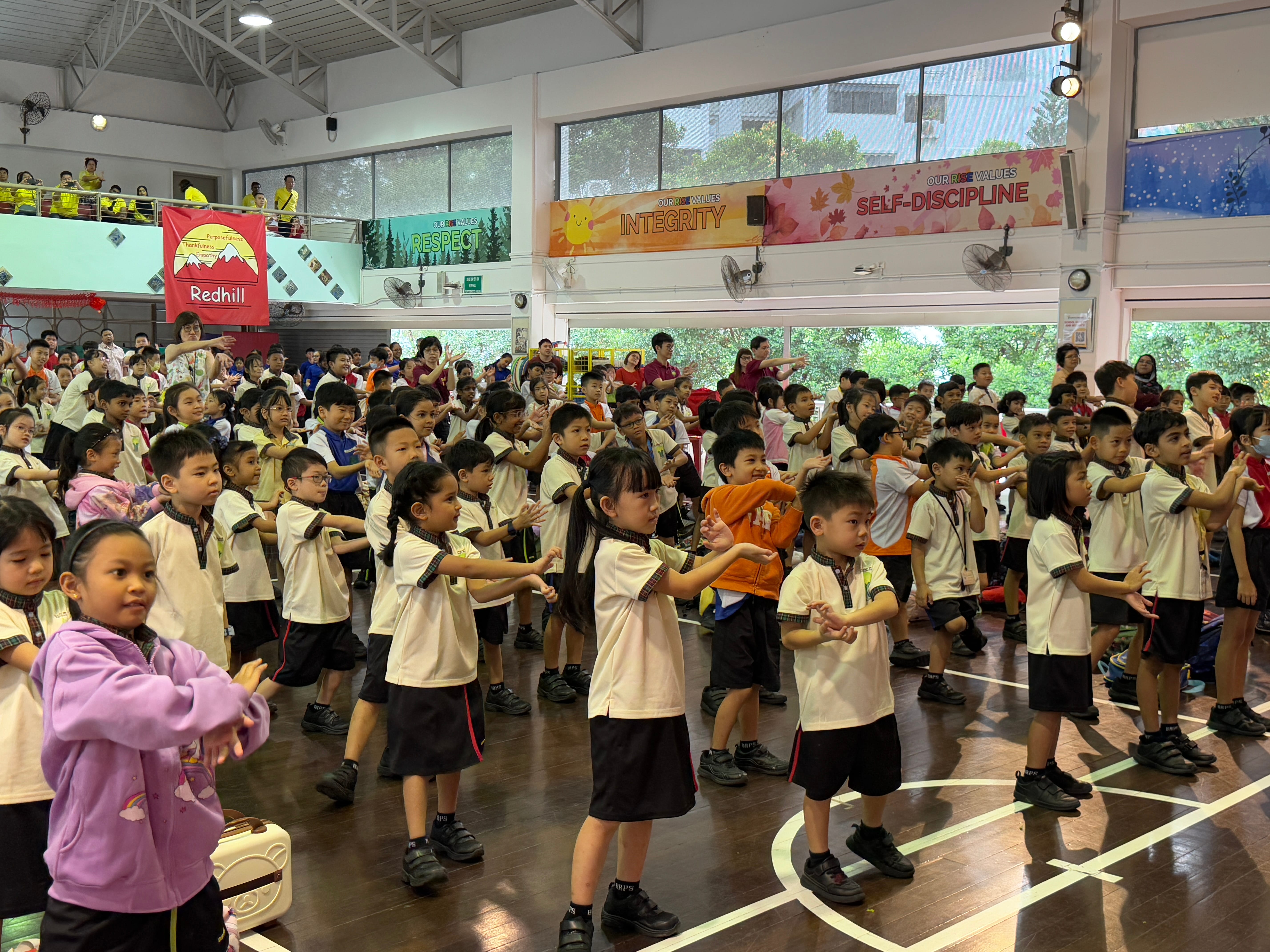 Schoolchildren in uniforms in a hall, arms out in unison, with banners and a balcony crowd above.