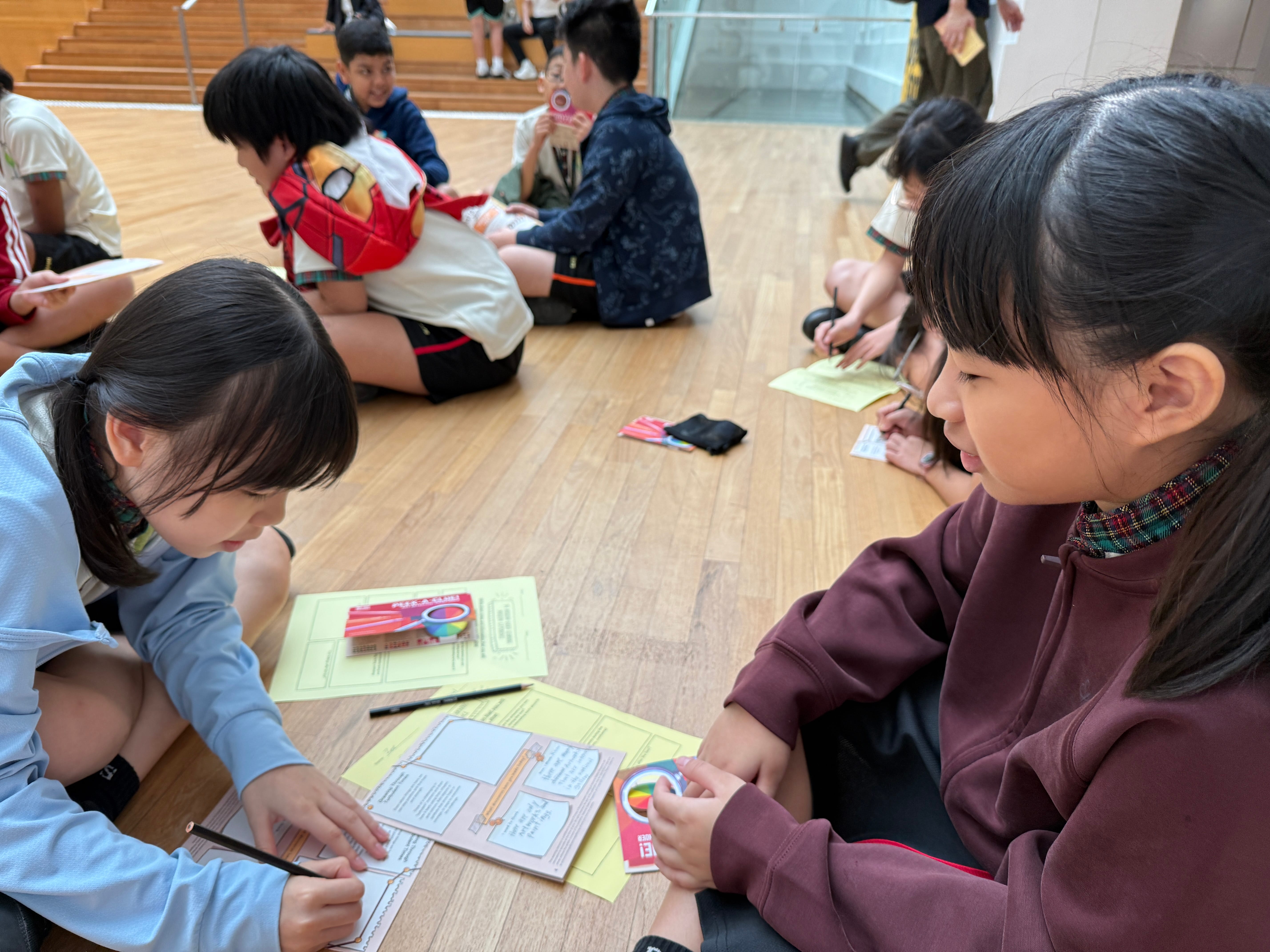 Children sit on a wooden floor in groups, writing on worksheets with pens and colored cards.