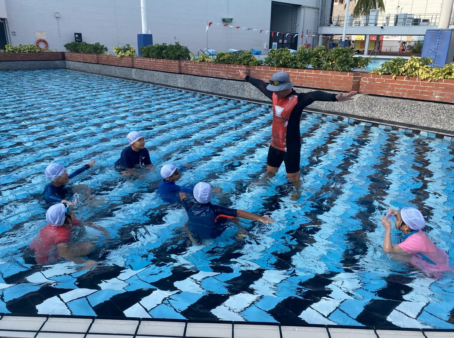 A swim instructor stands in a pool with arms outstretched, demonstrating technique to five children wearing white caps.