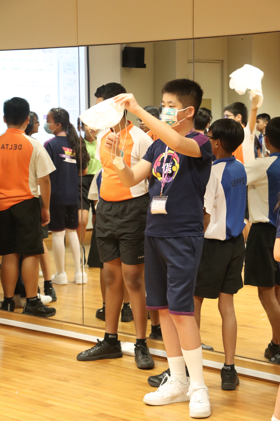 A masked boy in a navy shirt holds up a face mask in a mirrored room filled with other school children.