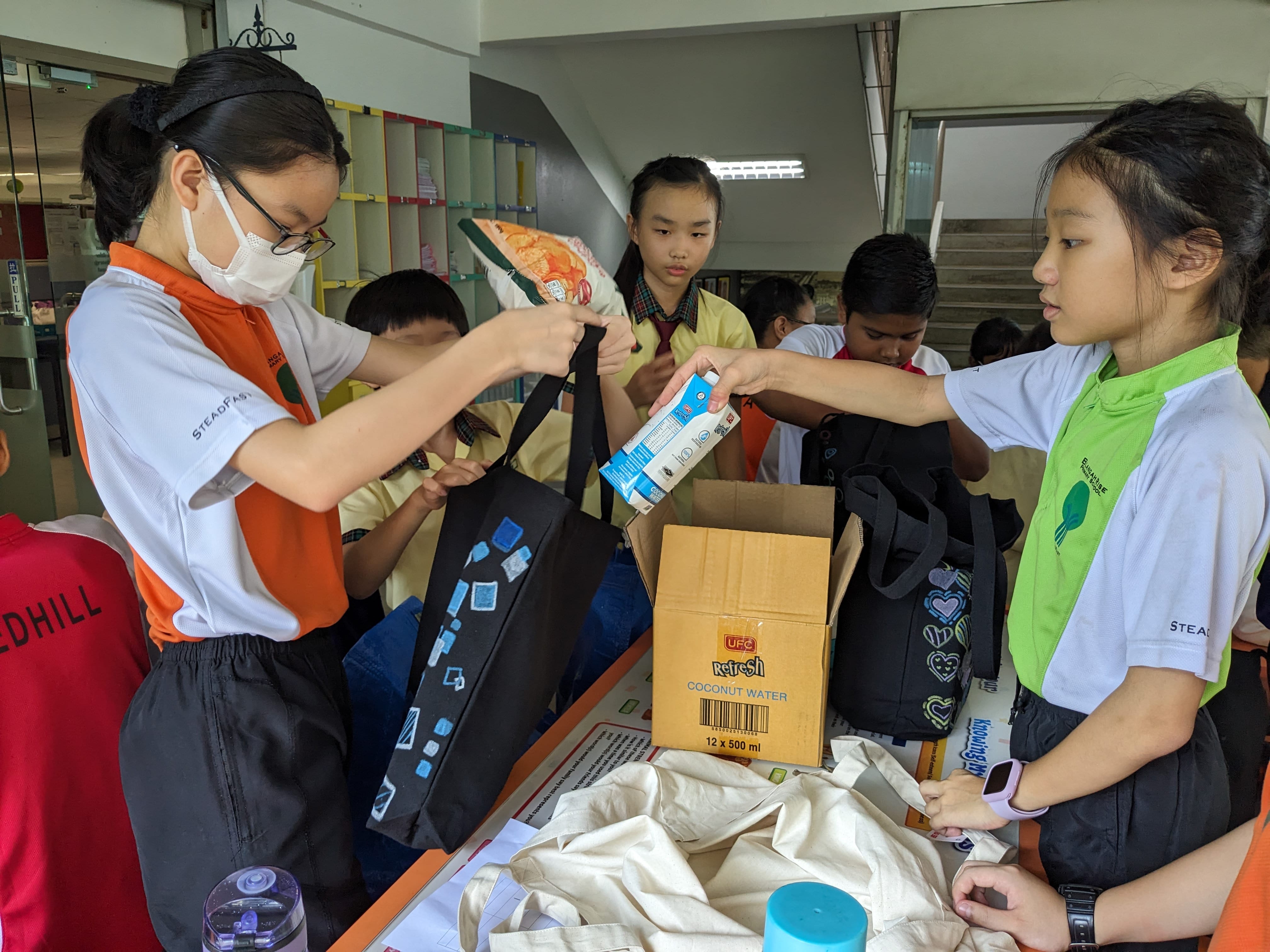 Students pack drink cartons and food into tote bags at a table, with an open box and stairs behind.