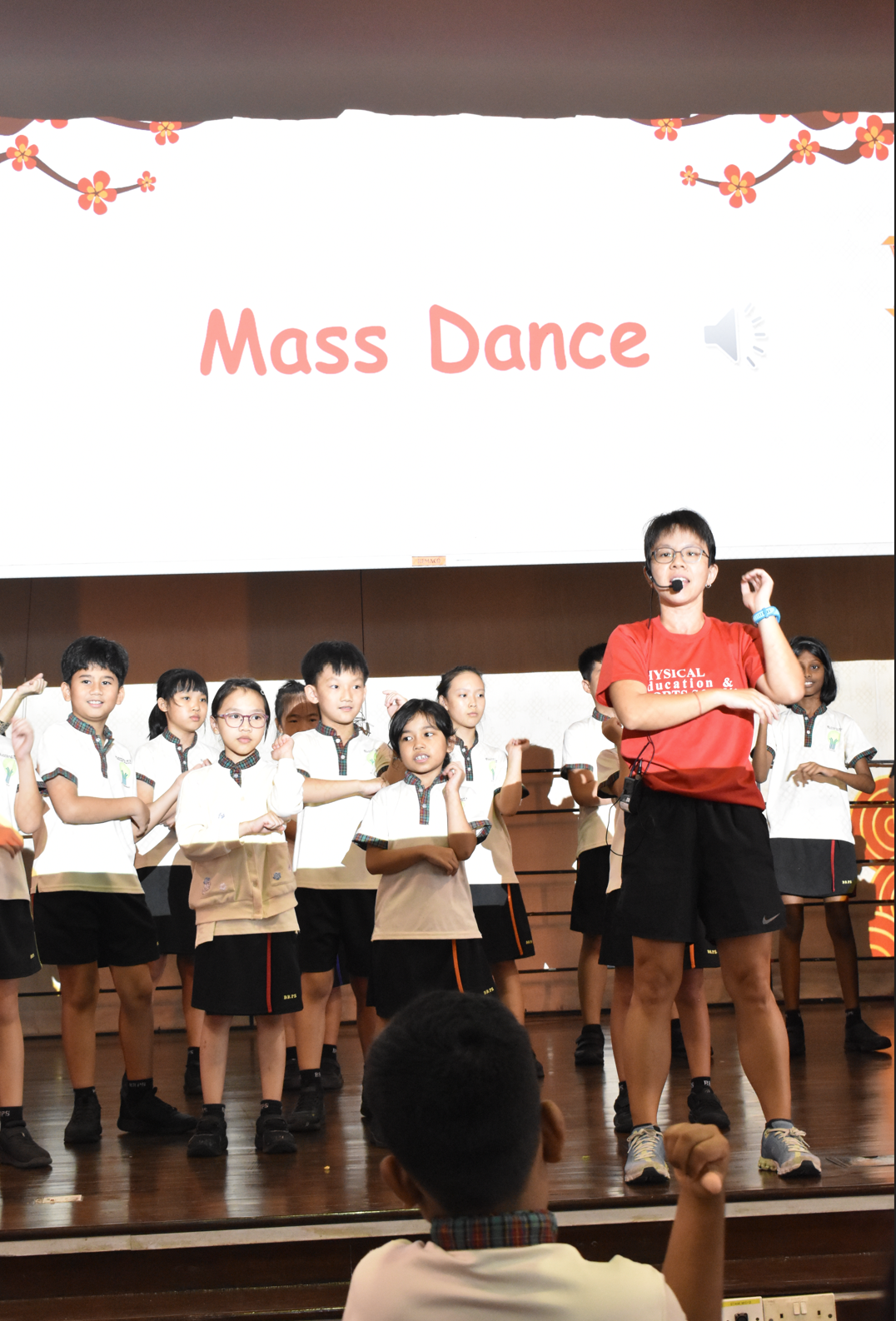 A teacher in red leads schoolchildren on stage in a Mass Dance activity, with a projection screen showing "Mass Dance" behind them.