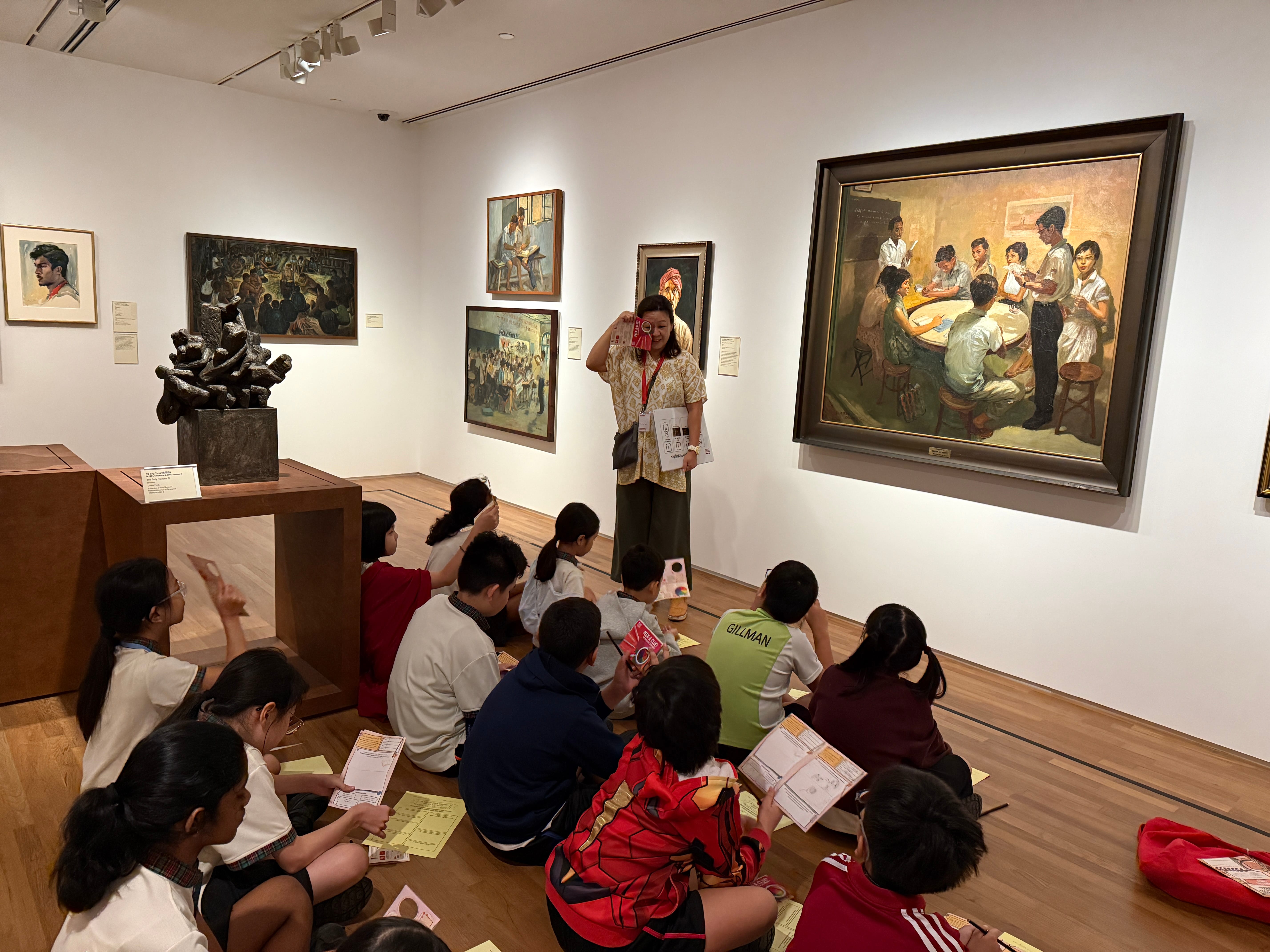 Teacher stands in art gallery as seated children sketch and look at paintings; dark sculpture on pedestal nearby.