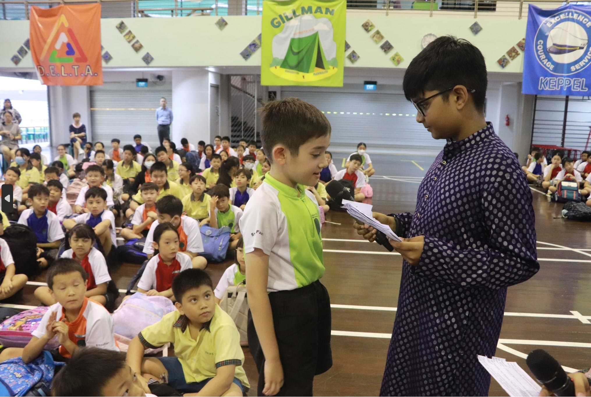 A student holds a microphone toward a young boy during a school assembly with many children seated on the floor.