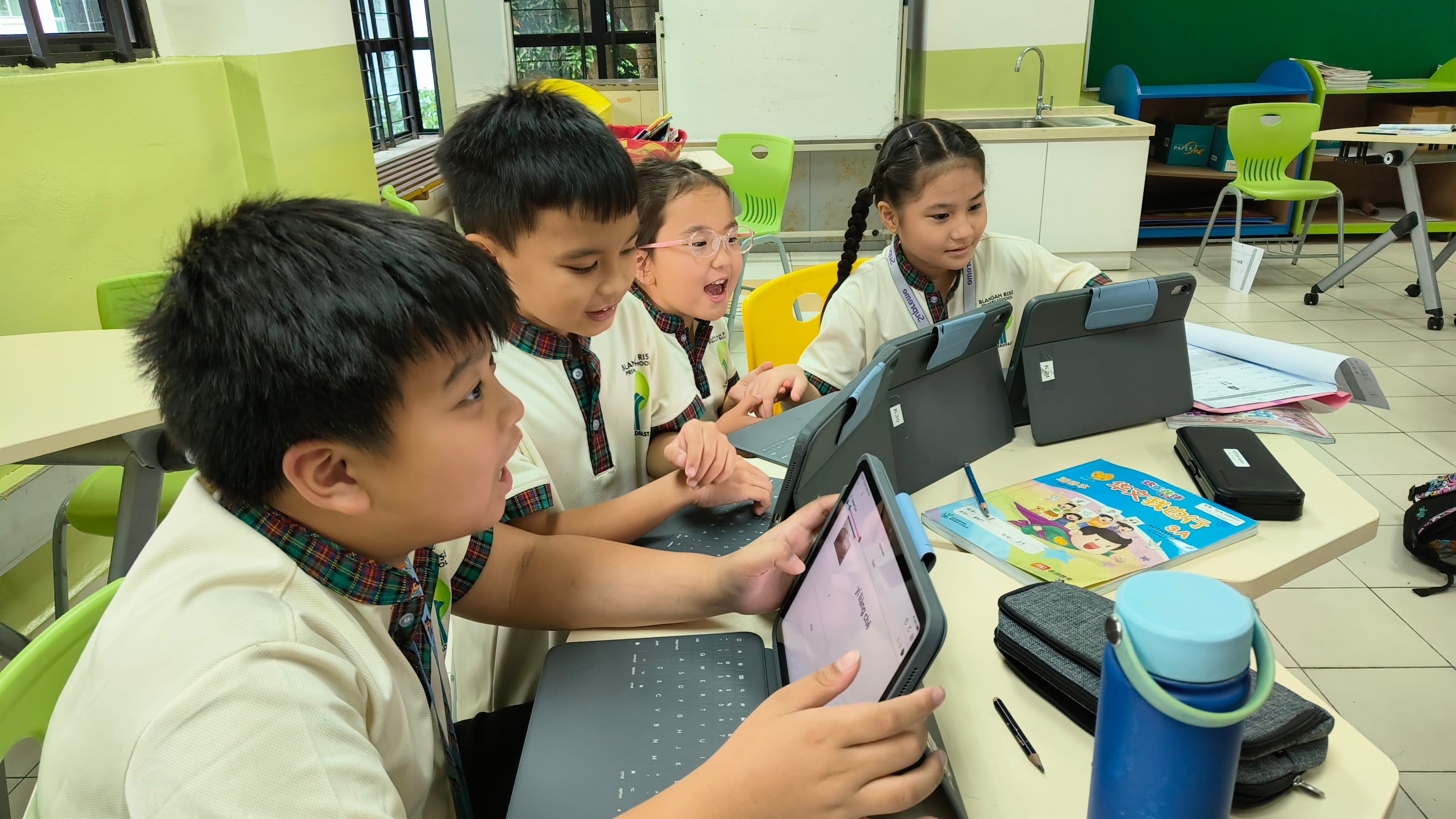 Four children in school uniforms sit at a table using tablets with keyboards in a classroom.