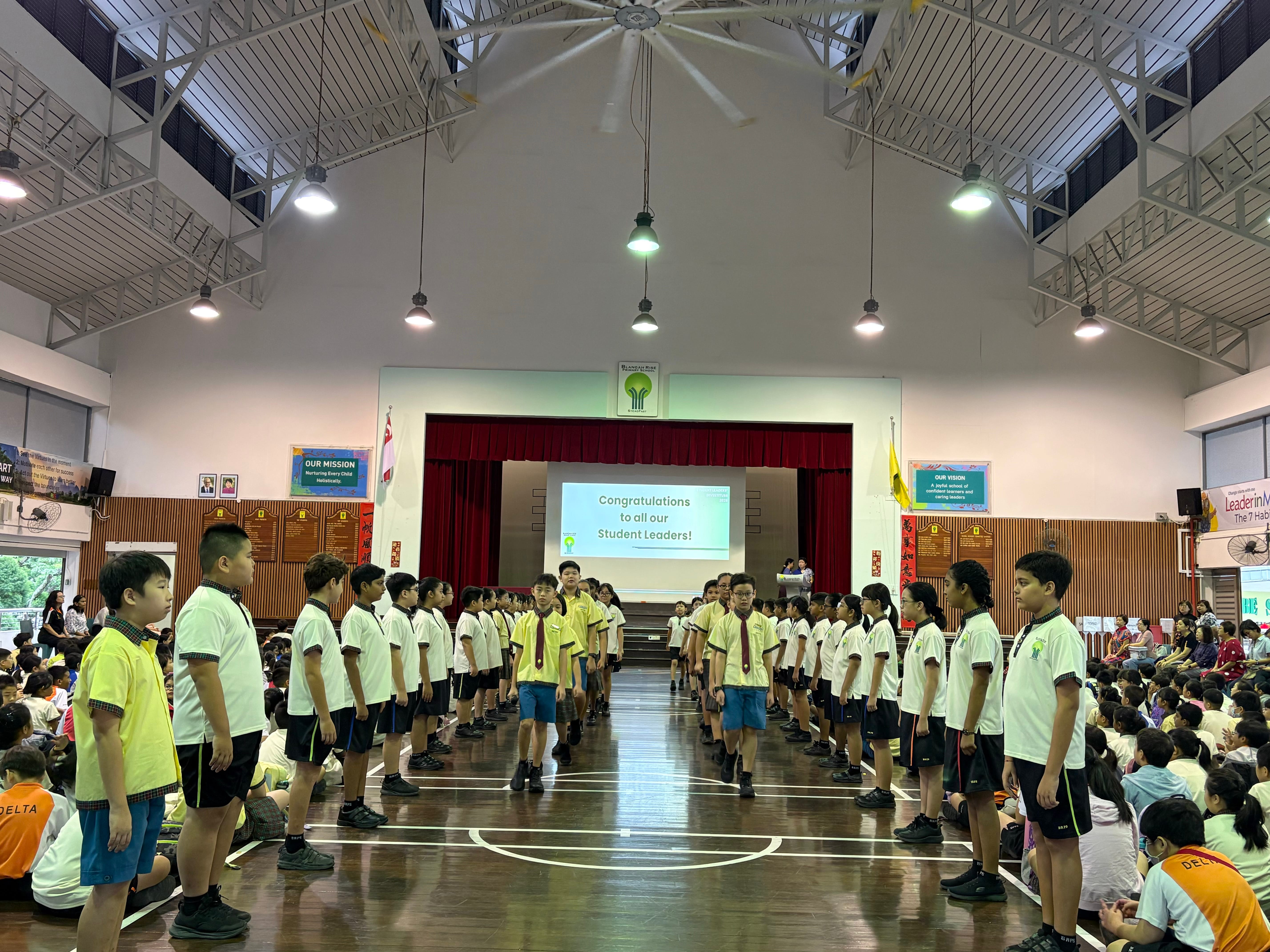 School assembly in a hall, students lined in two rows facing each other, stage screen reads congratulations.